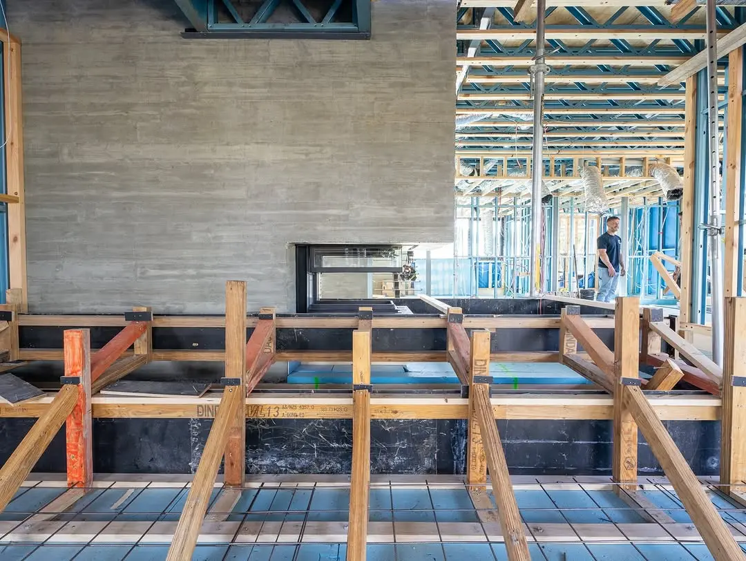 Interior view featuring a massive board-marked concrete wall, wooden formwork and bracing, and blue slab insulation with steel reinforcement in the foreground.