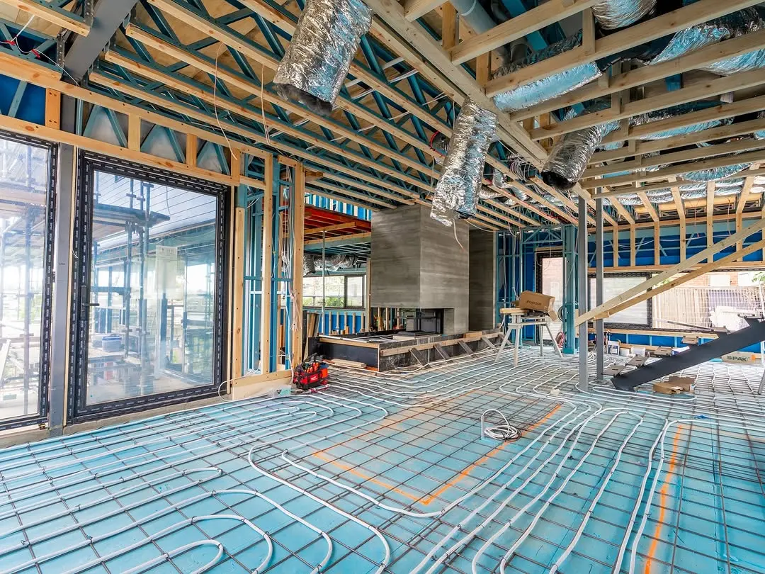 Wide interior view of the building under construction, showing a network of underfloor heating pipes laid over blue insulation, with exposed timber ceiling trusses and silver ducting above.