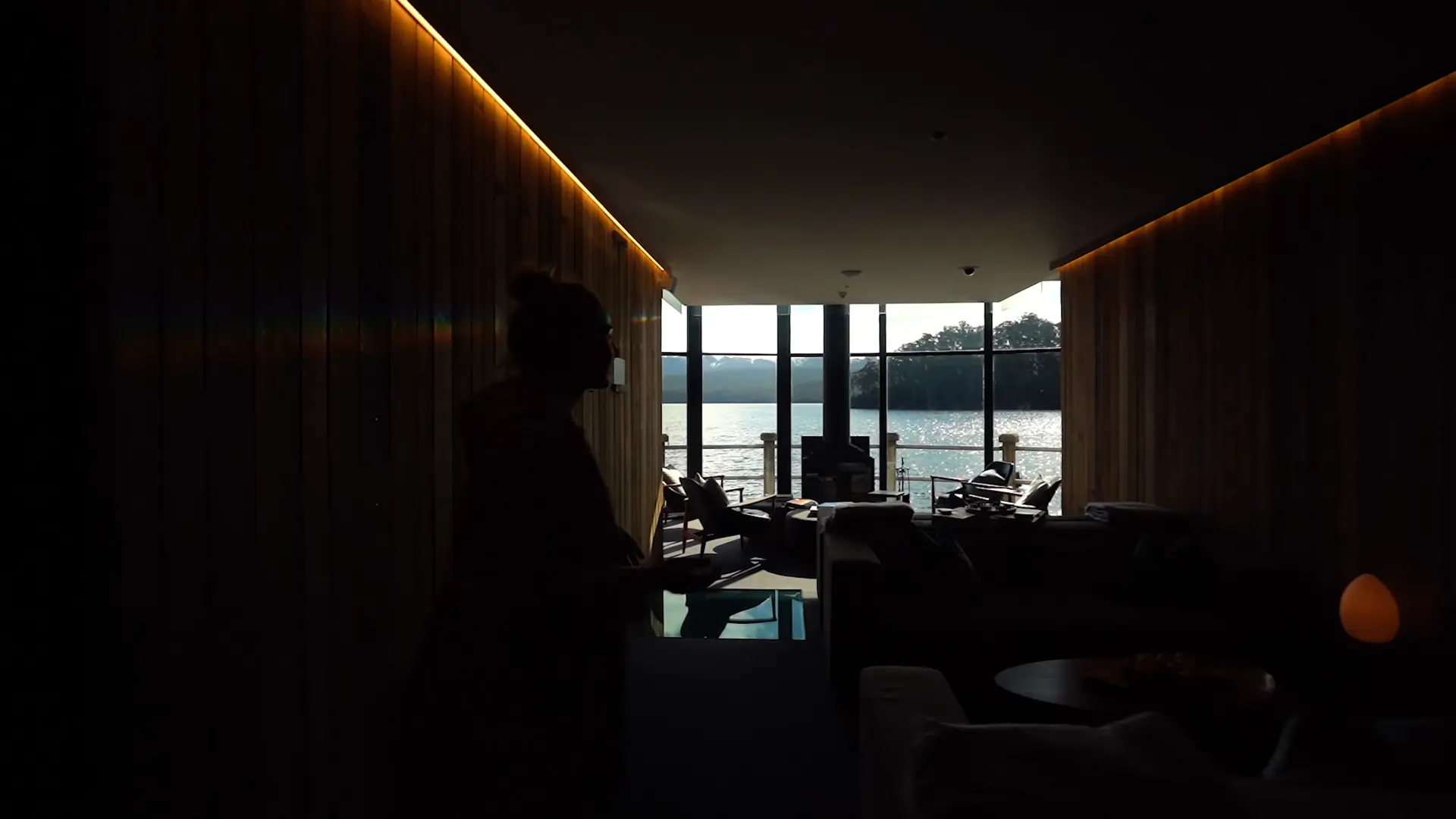 Video still of the interior lounge space at Pumphouse Point with floor-to-ceiling glazing facing Lake St Clair, timber-lined walls, and a fireplace, as Lucy Glade-Wright gestures toward the expansive landscape beyond.
