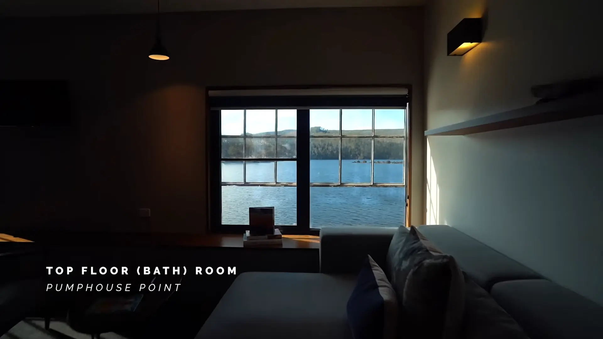 Video still of the top floor bedroom at Pumphouse Point overlooking Lake St Clair, with a large grid-framed window capturing expansive water views, minimal furnishings, and soft ambient lighting.