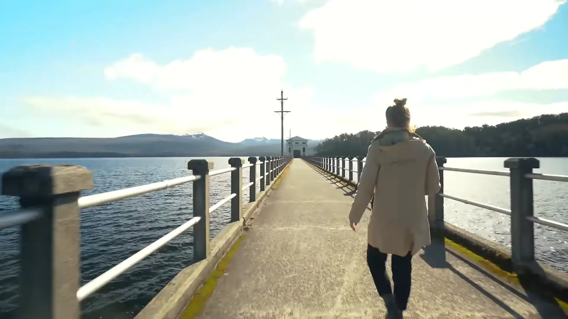 Lucy walks along the narrow concrete jetty toward the historic pumphouse, surrounded by Lake St Clair and distant mountain ranges under the bright Tasmanian sun.