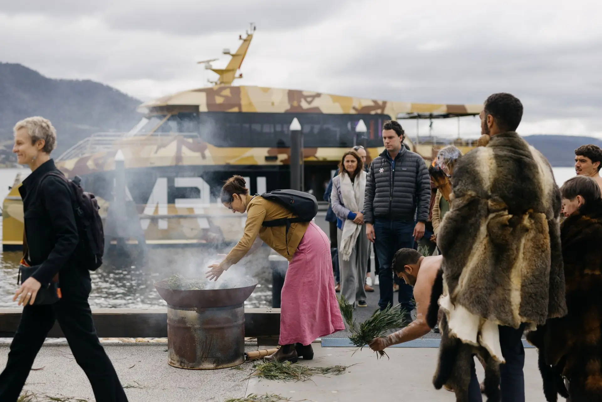 A Smoking Ceremony at Brooke Street Pier in Hobart, with participants gathered around a fire pit in front of a camouflaged MONA ferry.