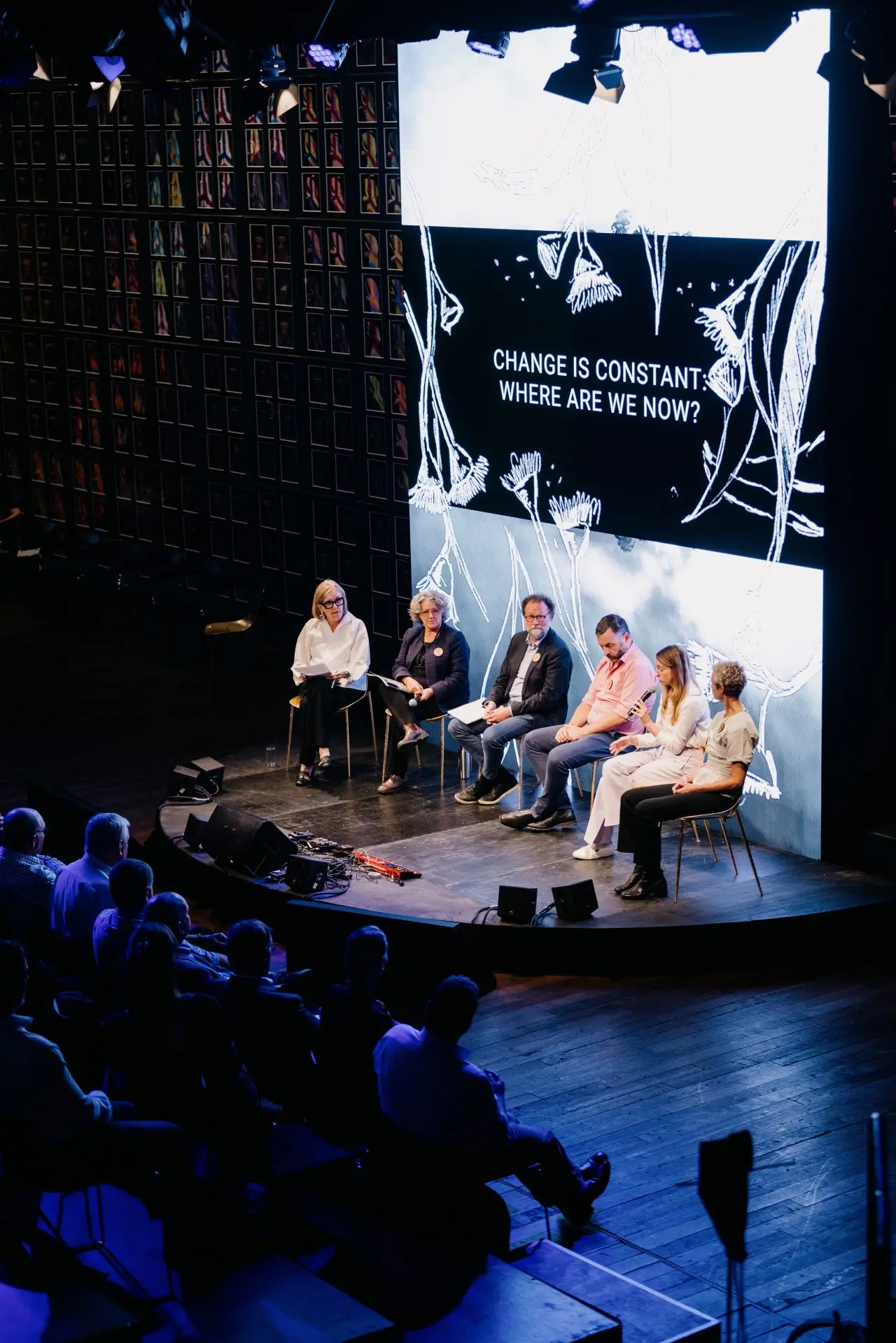 A panel of experts sits on a theater stage in front of a large screen reading "CHANGE IS CONSTANT: WHERE ARE WE NOW?", addressing an audience.