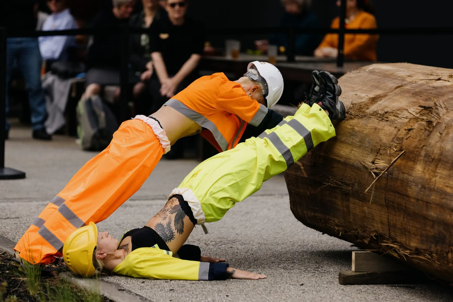 wo performers in high-visibility workwear and hard hats engage in an acrobatic physical performance involving a large log.