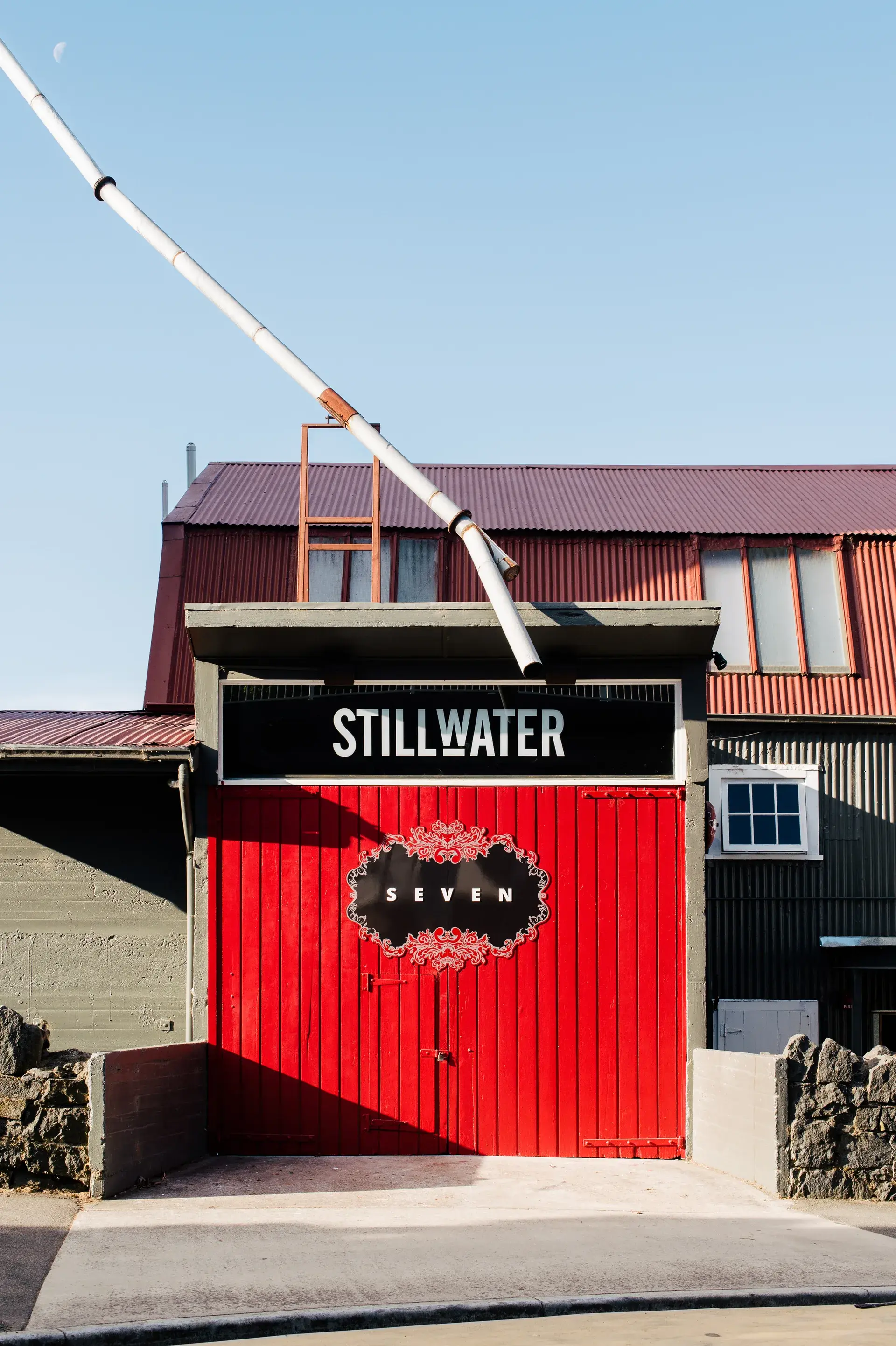 Large vibrant red doors with a black "SEVEN" emblem mark the entrance to "STILLWATER". The building features grey corrugated metal walls and a deep red roof, with a long white pole extending diagonally across the clear blue sky.