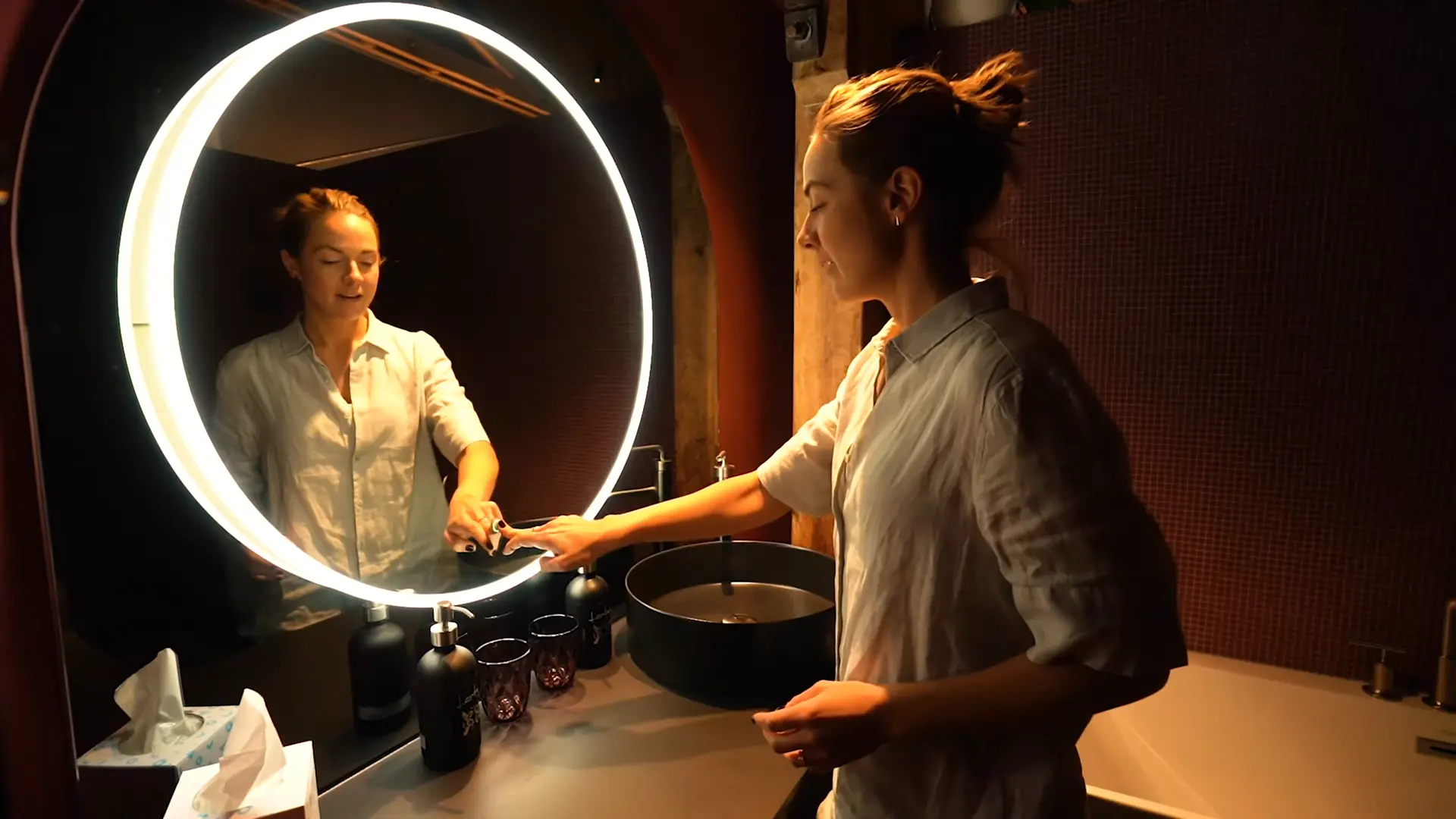 Lucy Glad-Wright using a circular backlit mirror in a warmly lit ensuite bathroom, lined in red tiles.