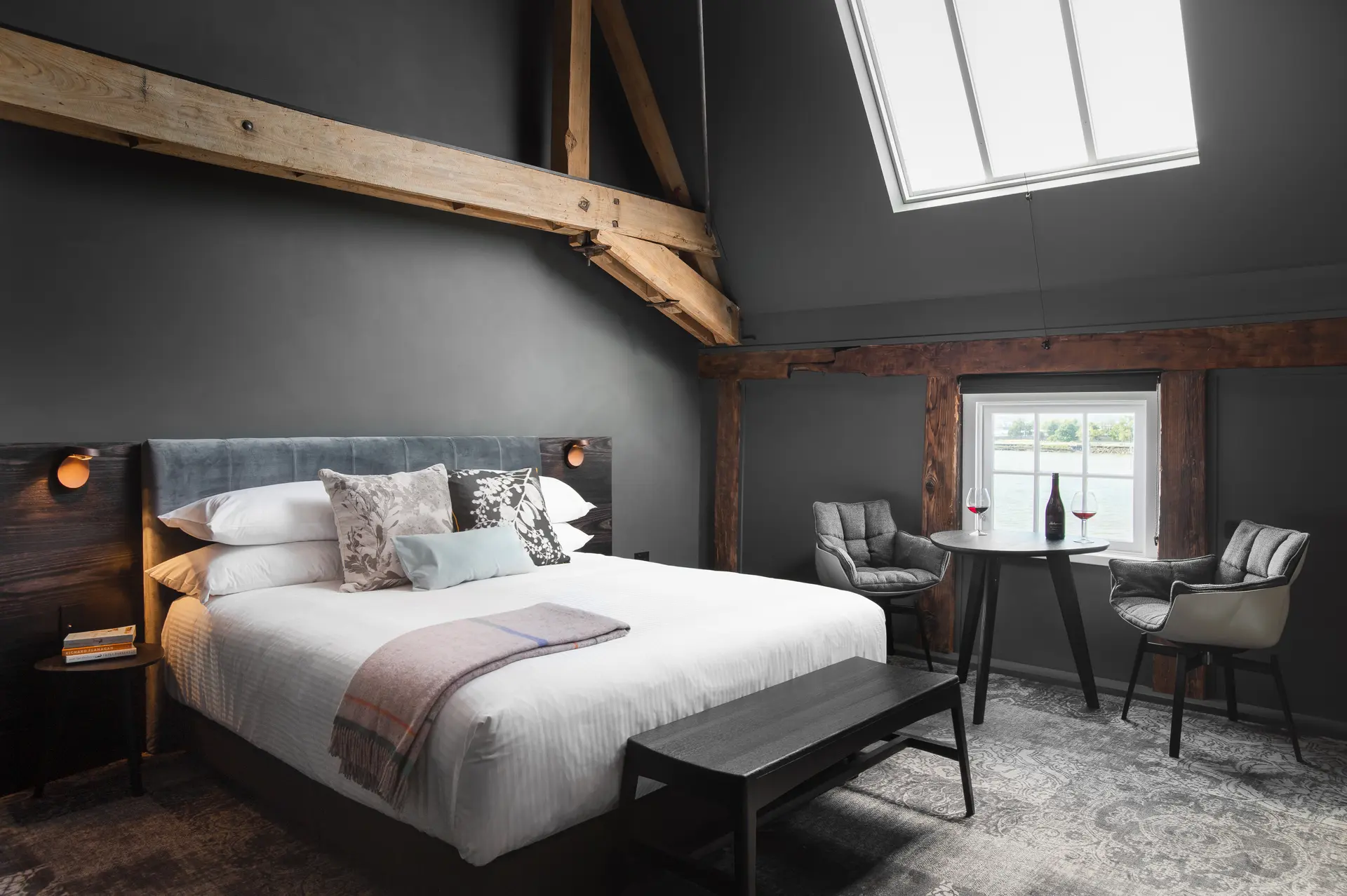 This industrial-style bedroom features exposed timber beams across a grey ceiling and dark grey walls. A small seating area with two grey chairs and a round table is positioned by windows framed in natural wood, while the bed is accented with a grey velvet headboard.