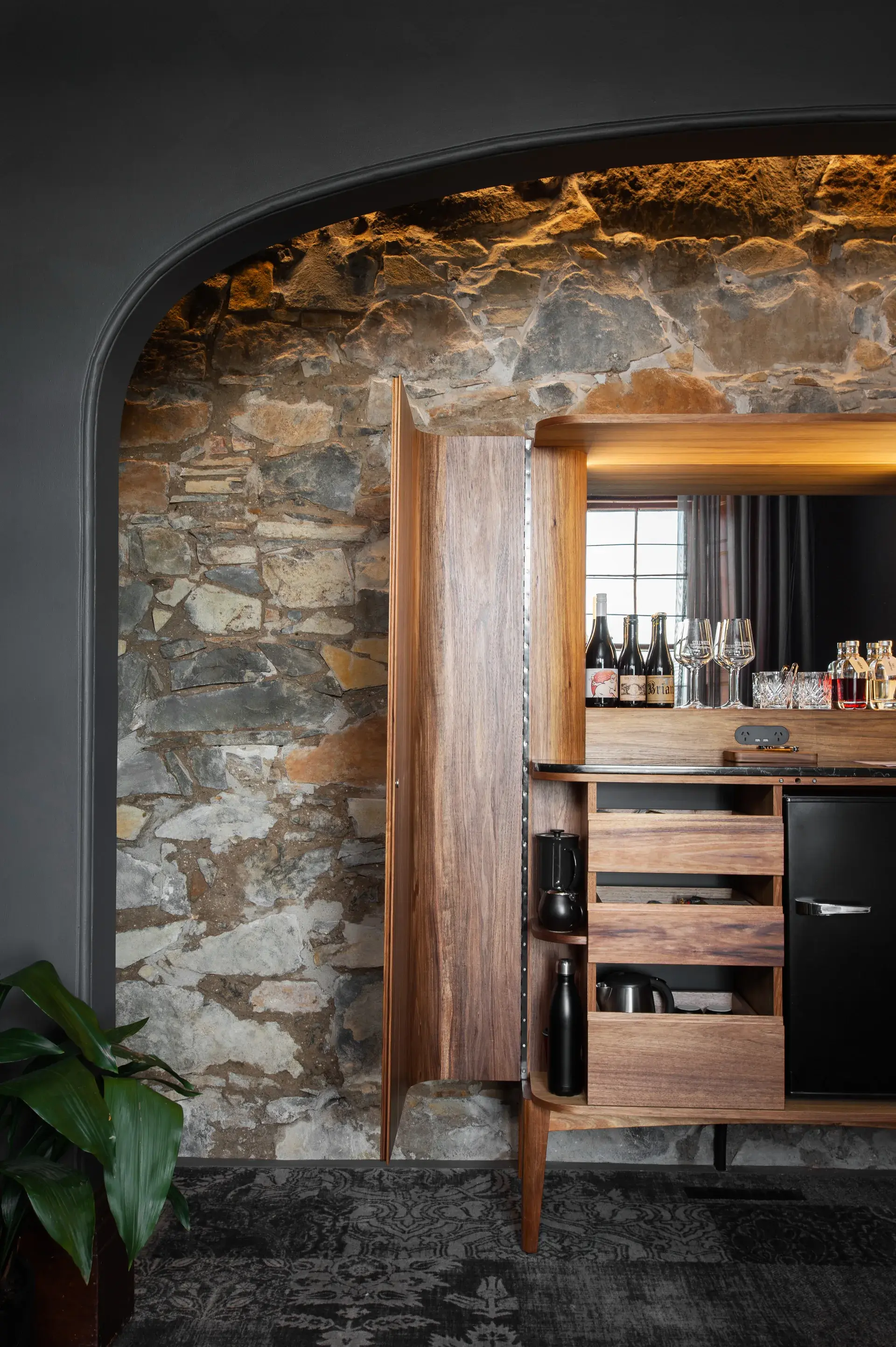 A wooden bar cabinet features a black marble top and a mirrored backsplash reflecting glassware. It includes a small black refrigerator and open drawers filled with snacks and drinks.