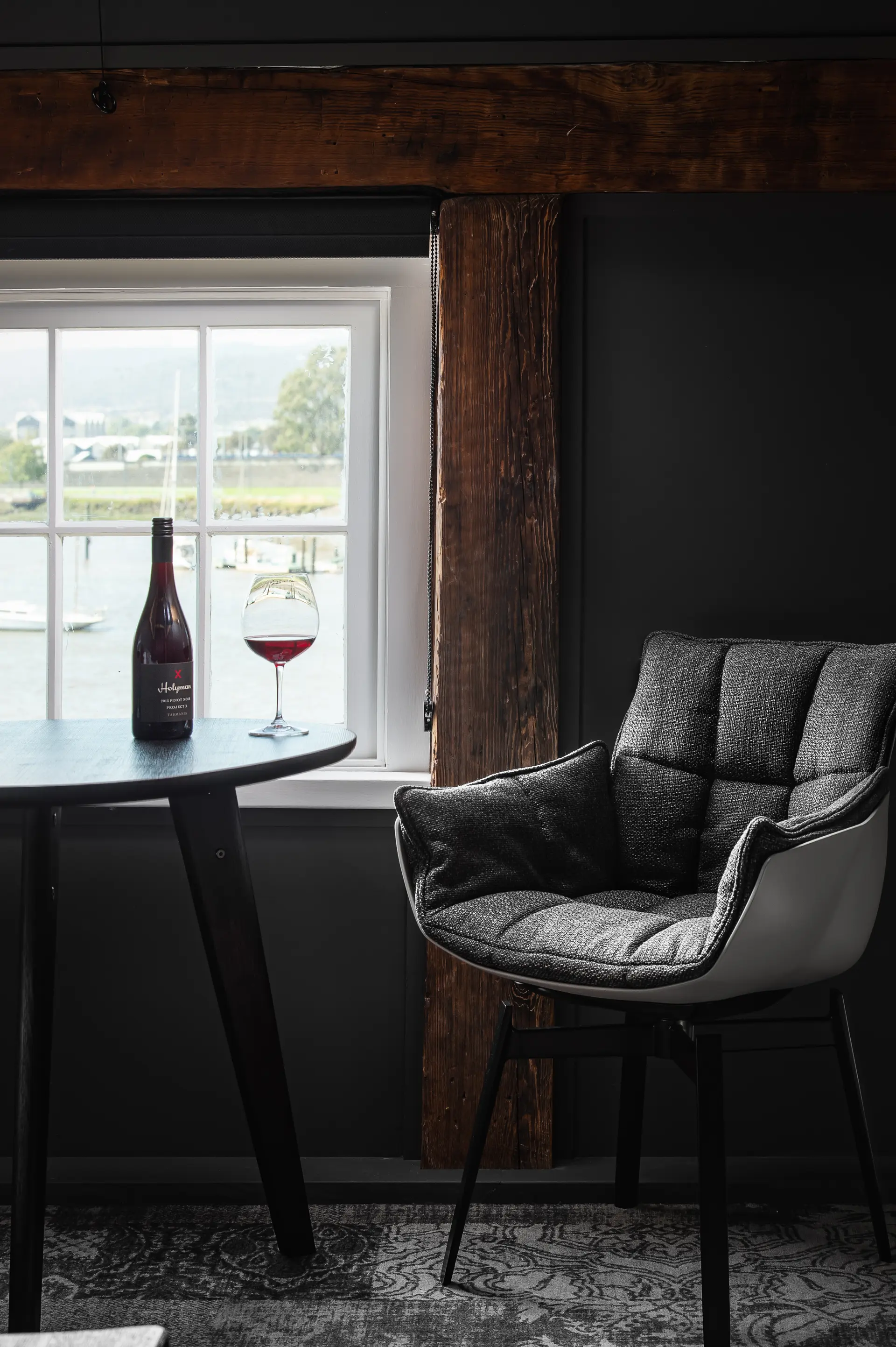 A sitting area in one of the Stillwater Seven suites, featuring an original Oregon pine beam, dark walls, a round table with a glass of red wine overlooking the Tamar River, and a grey upholstered chair.