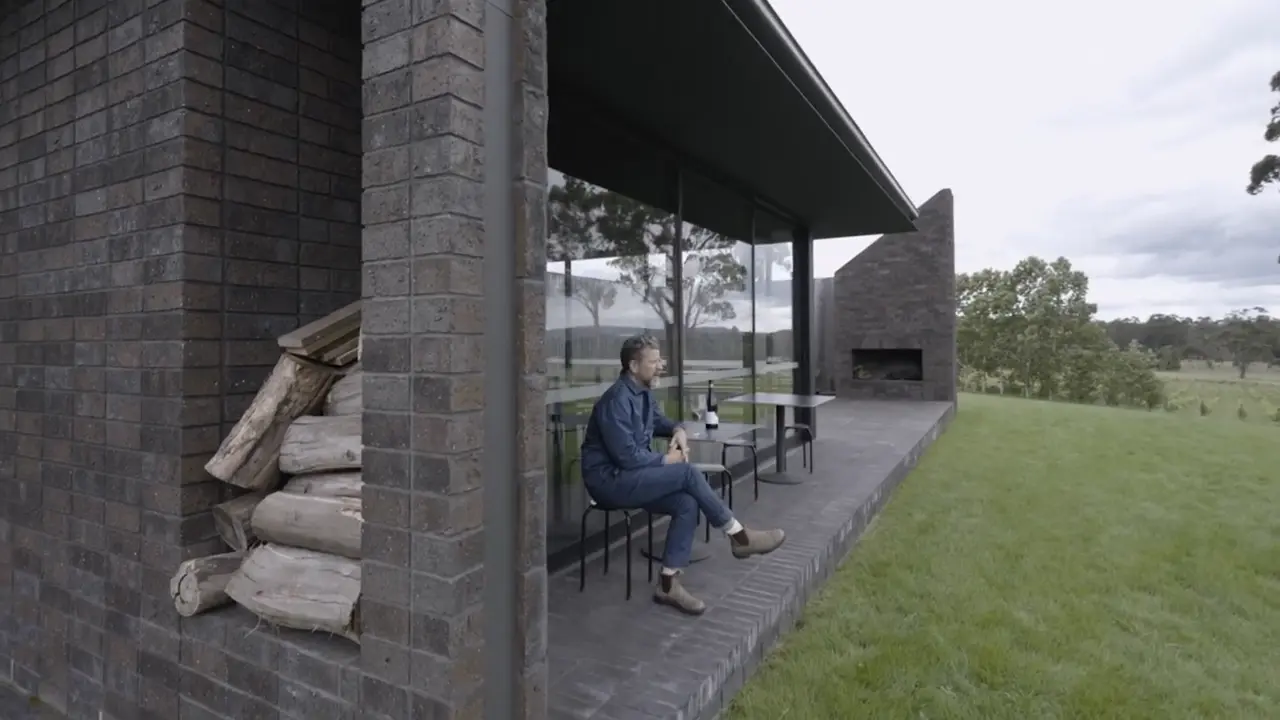 A man in denim and boots sits on the terrace of a dark-brick vineyard pavilion, featuring a built-in wood storage niche and an outdoor fireplace, overlooking a vast Tasmanian rural landscape.