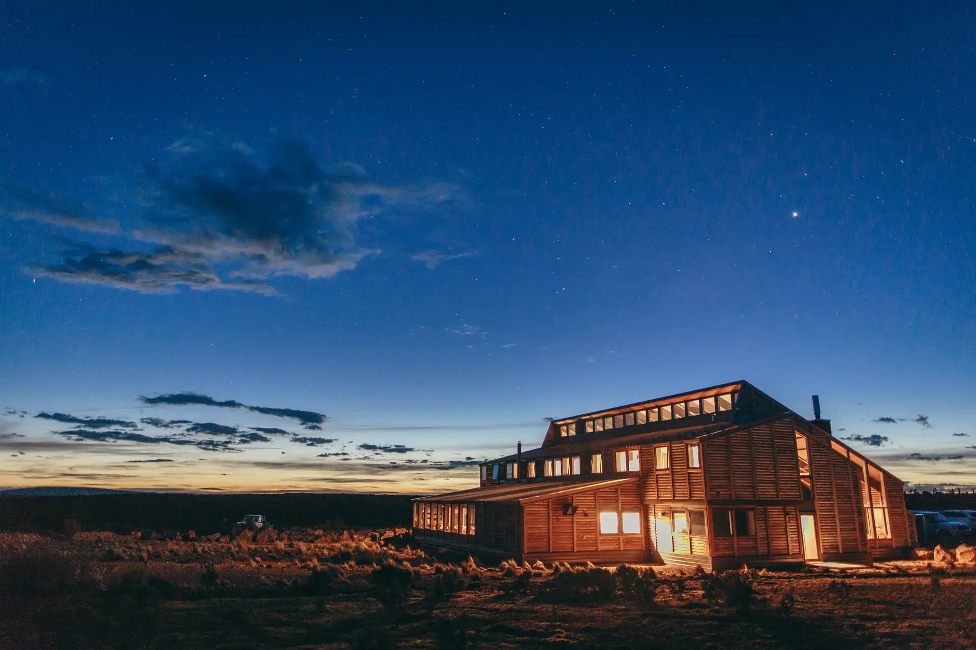 This large, multi-story timber lodge features a slanted roofline and glowing windows, set against a deep blue twilight sky filled with stars. The building is constructed with horizontal wood siding and includes a long row of clerestory windows along the top level.