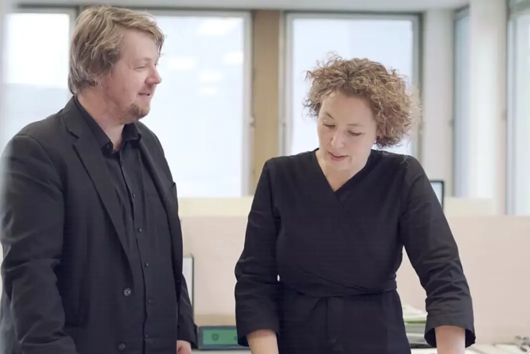 Cumulus Co-Founders and Directors Peter Walker and Kylee Scott standing together in a bright studio workspace, reviewing drawings on a desk and engaged in discussion, with large windows and a light-filled office environment in the background.