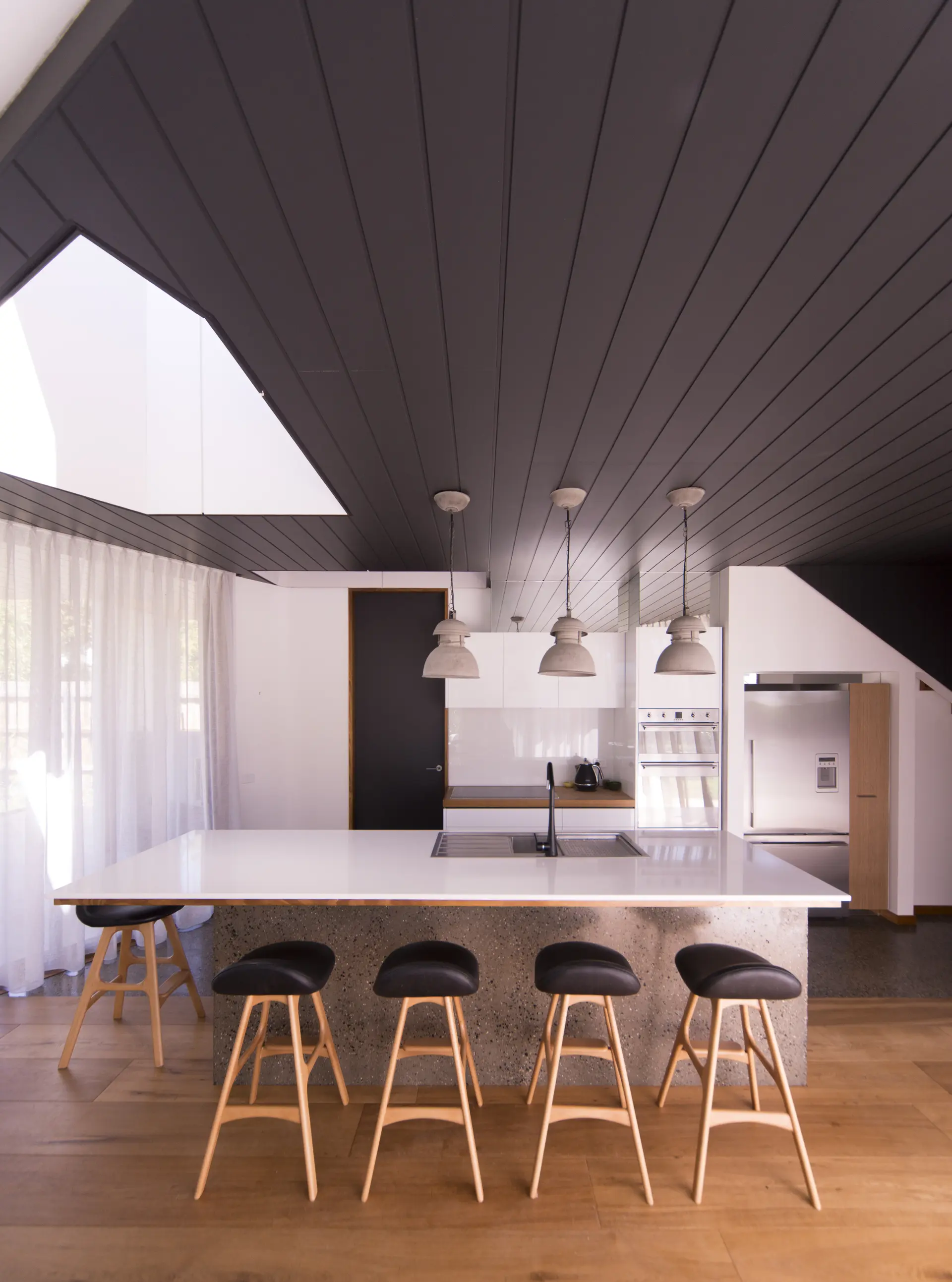 Inside, the kitchen and dining area is organised beneath a central skylight, bringing daylight into the centre of the plan. A polished concrete island is paired with white joinery, timber flooring, and a timber-lined ceiling.