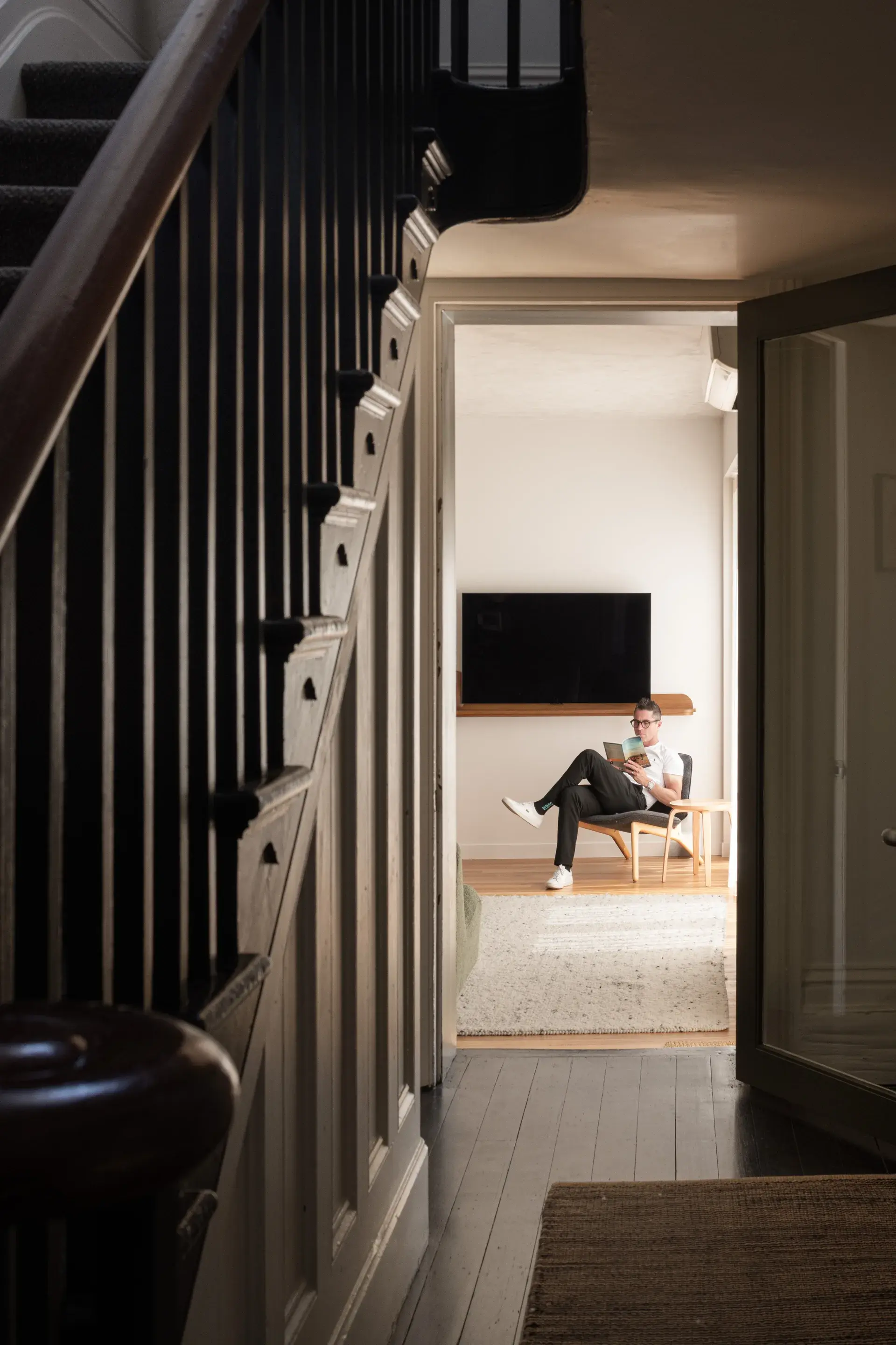A dark-stained wooden staircase with a prominent bannister frames the foreground of this residential hallway. Through an open doorway, a person sits reading in a bright room featuring a wall-mounted television and minimalist furniture.
