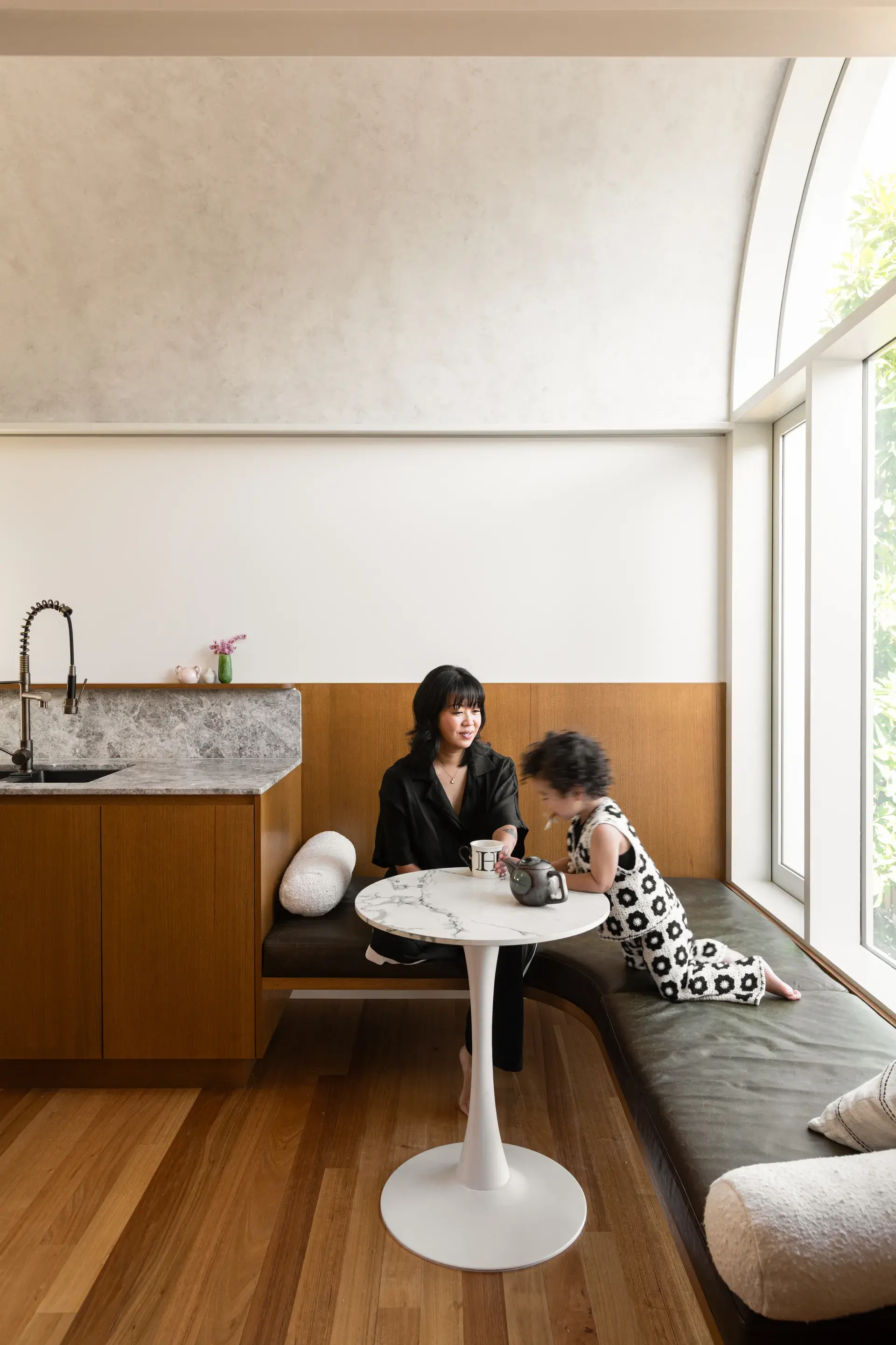 This bright, modern kitchen and dining nook features a white marble tulip table paired with a curved, dark leather banquette. A large arched window floods the space with light, highlighting the wooden cabinetry, stone countertops, and minimalist white walls.