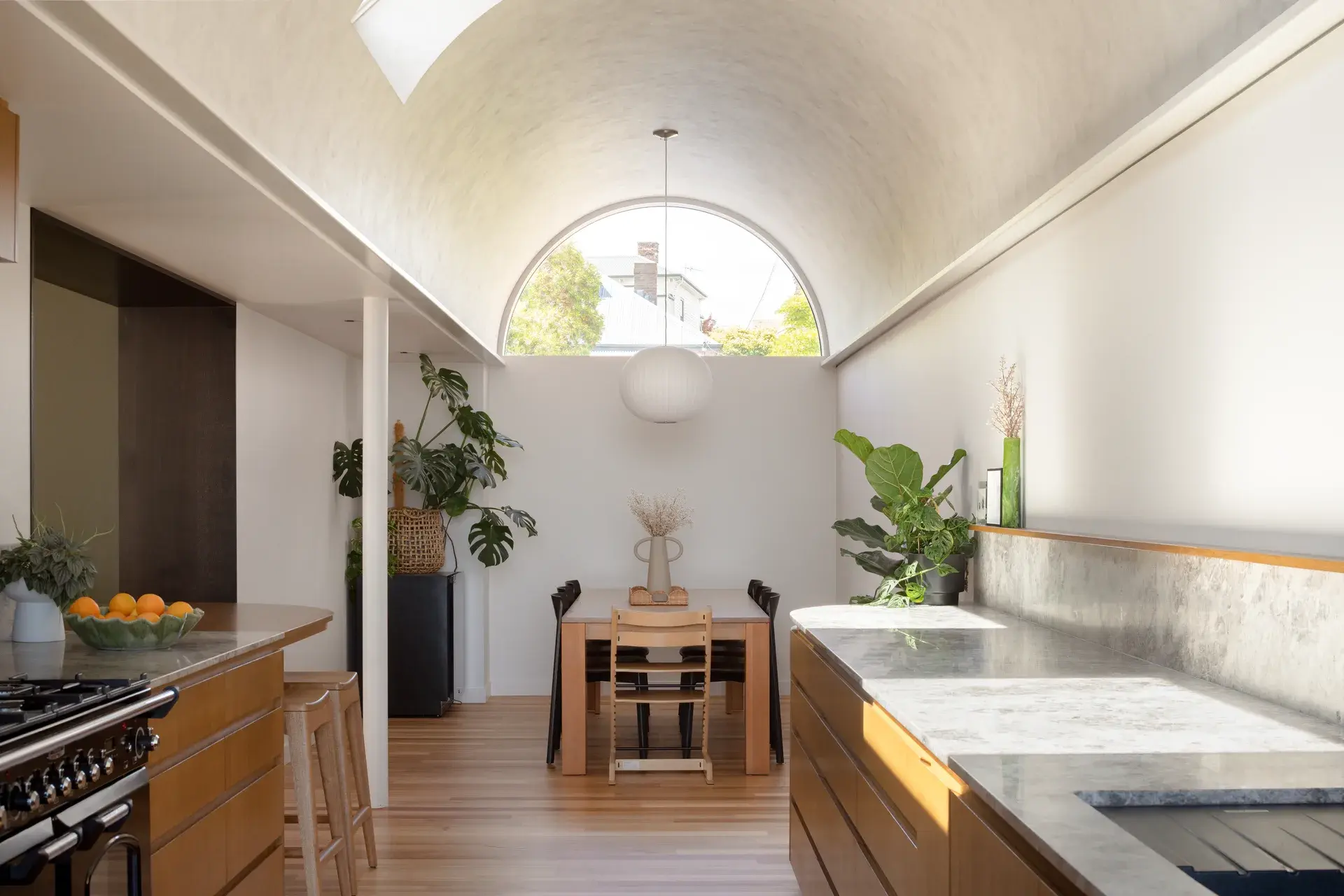 This long, open-plan space features a striking barrel-vaulted ceiling and a large arched window. The design includes sleek wood cabinetry with stone countertops and a dining table set beneath a spherical pendant light.