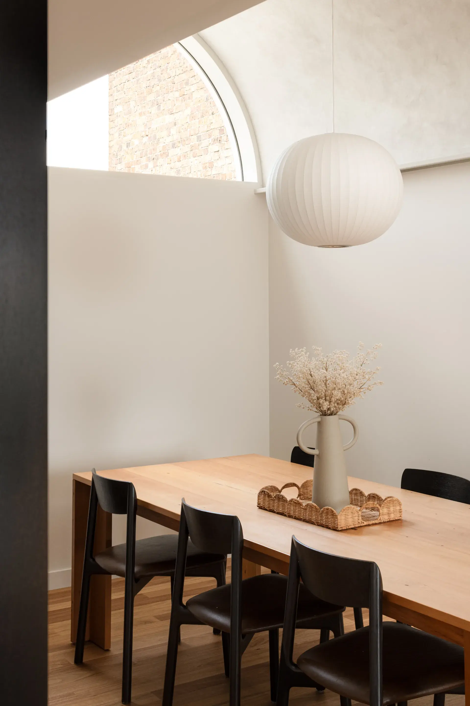 This minimalist dining space features a light wood table paired with black modern chairs and a large, spherical paper lantern. A tall, sculptural vase with dried flowers sits on a woven tray, positioned beneath a large arched window that shows a brick exterior.