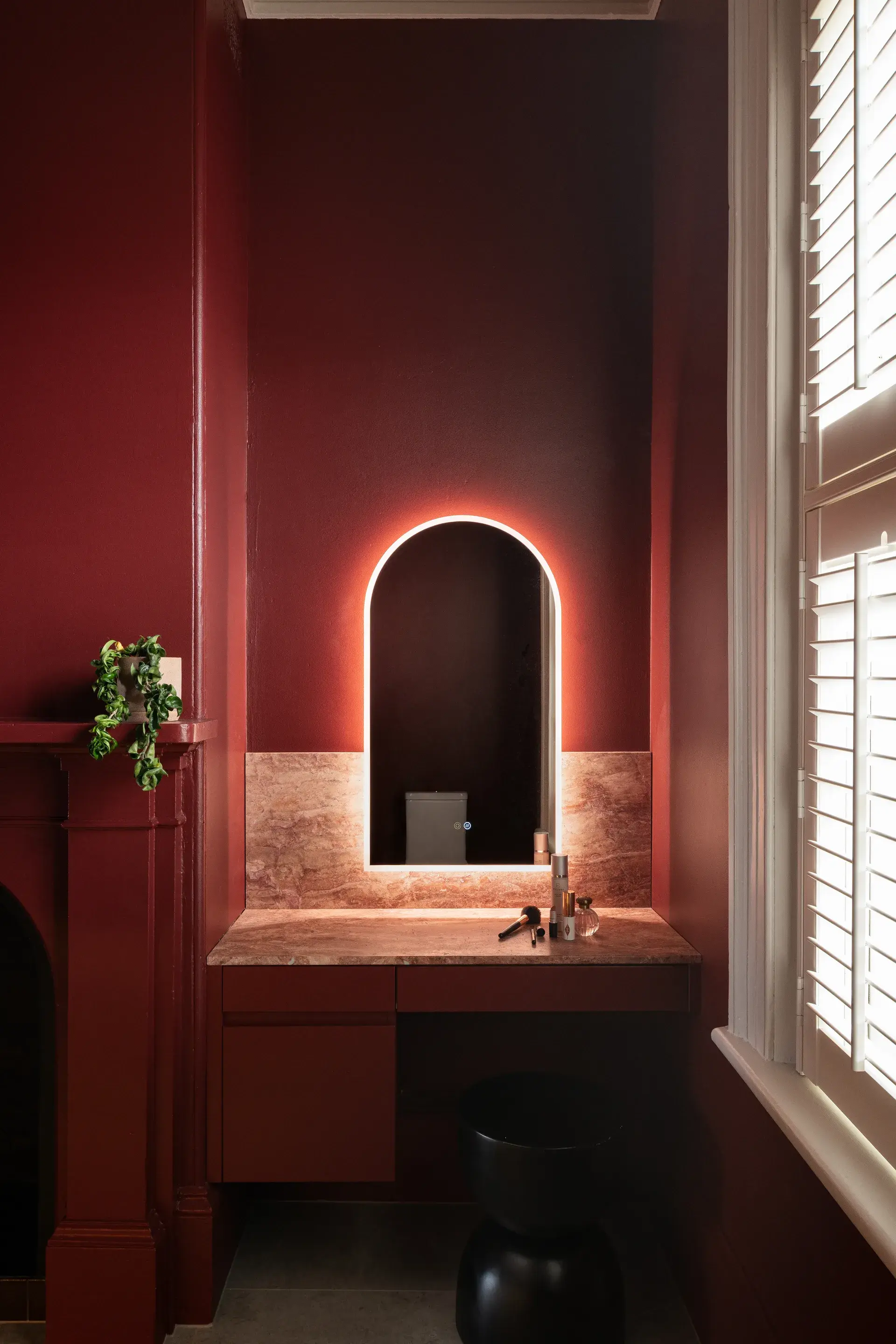 This elegant vanity area features deep burgundy walls and an arched mirror backlit with a soft orange glow. The mirror is set above a pink marble countertop and backsplash, with a matching burgundy desk and a sculptural black stool below.