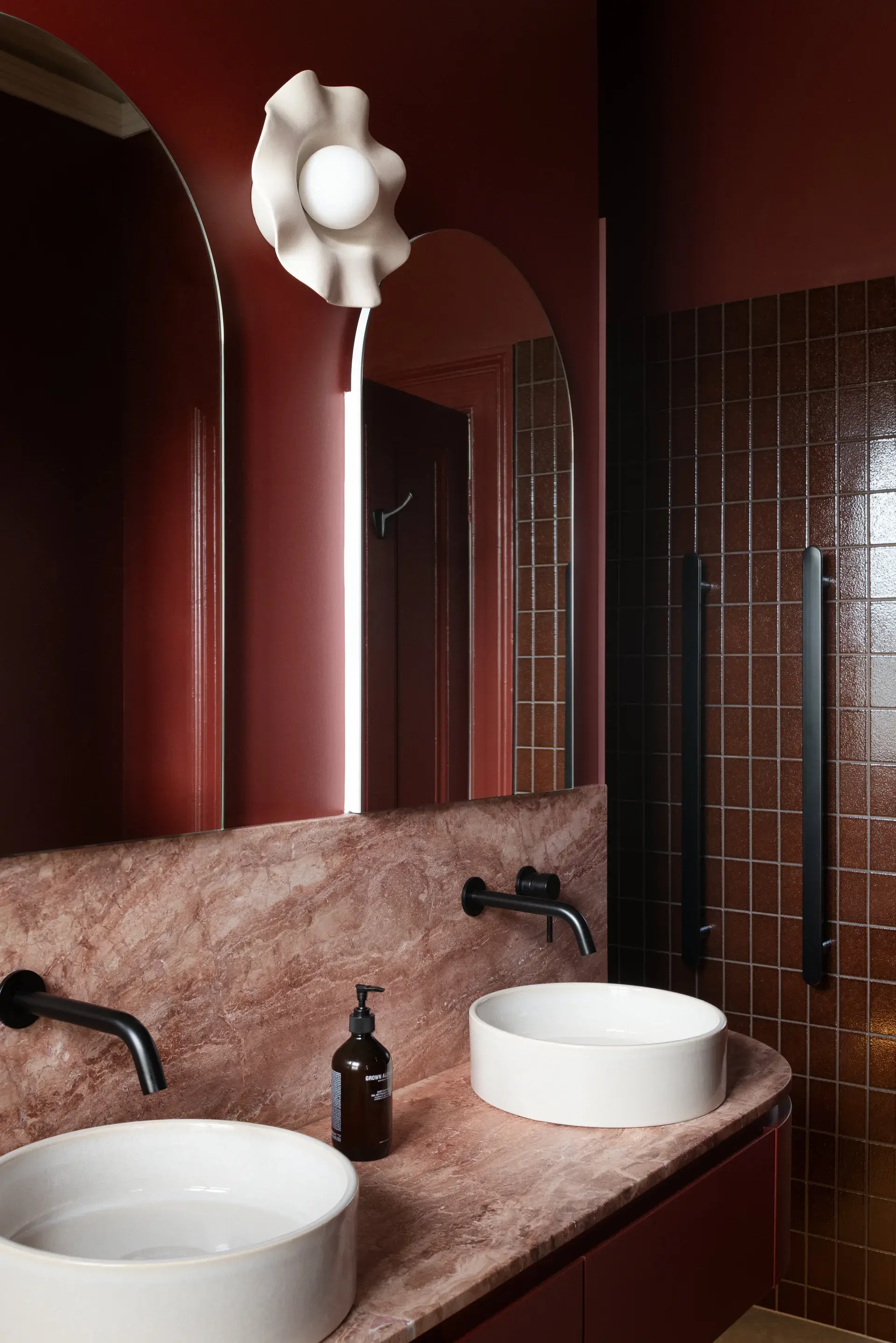 This sophisticated bathroom features dual white vessel sinks set upon a pink marble countertop and backsplash. Against deep burgundy walls, two arched mirrors are mounted beneath a sculptural, wave-like wall light, while dark vertical tiles line the adjacent shower area.