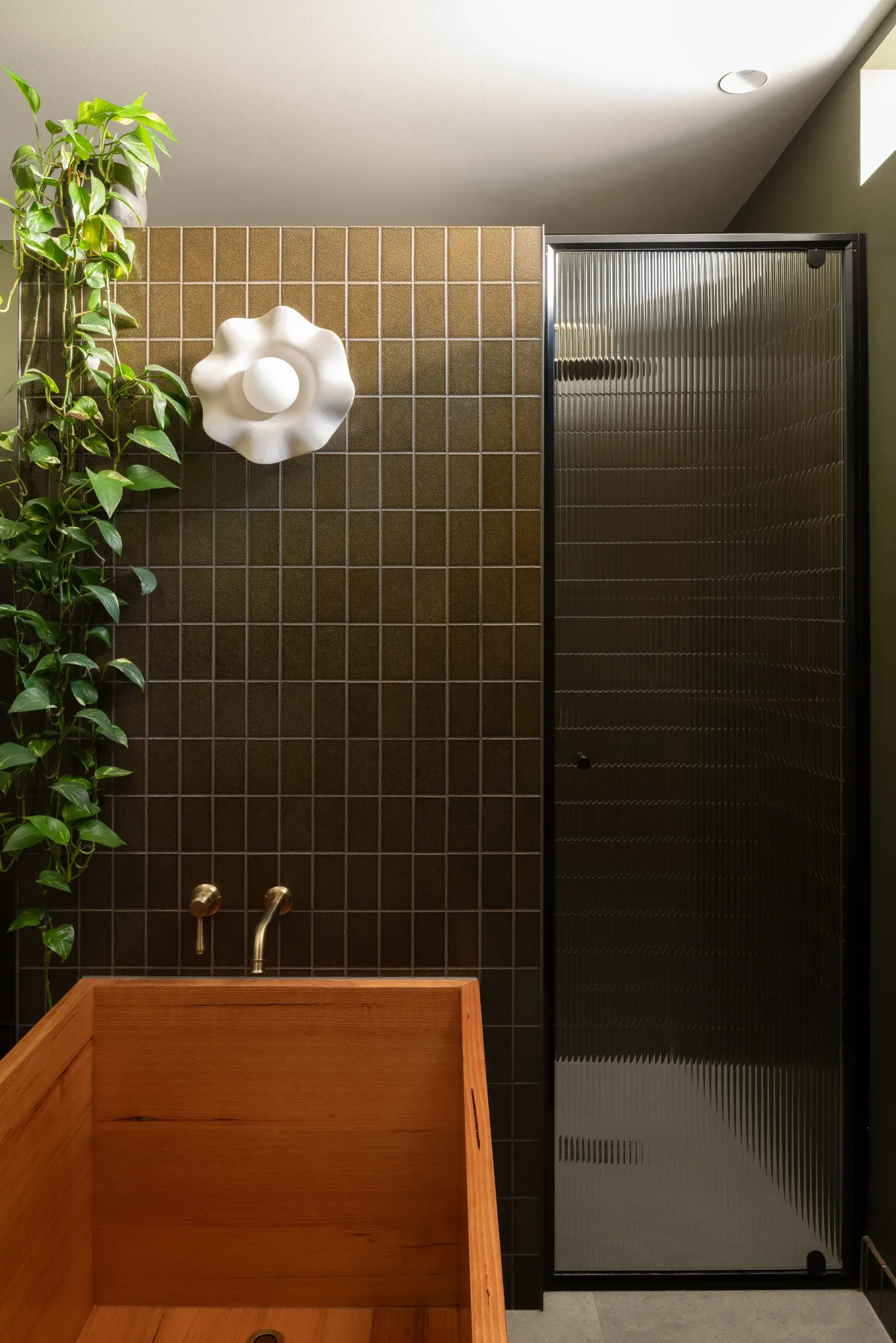 This modern bathroom features a hand-crafted wooden bathtub positioned in the foreground. Behind it, a dark-tiled wall is decorated with a climbing green plant and a sculptural white wall light, while a fluted glass shower door stands to the right.