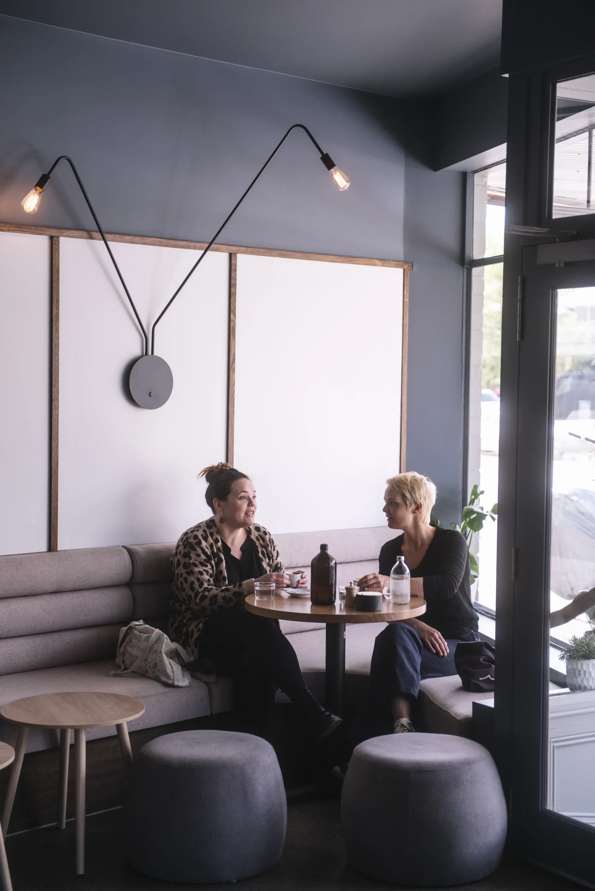 Two people occupy an intimate window alcove featuring a light grey couch that wraps around a timber table, lit by a playful lamp overhead and set against a wood-trimmed wall dividing dark grey and white finishes..