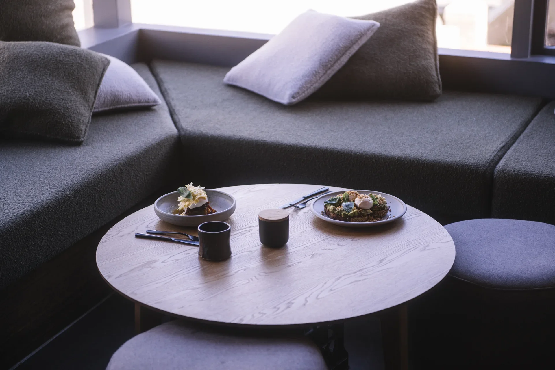 A minimalist seating space features a circular timber table and window bench seating.
