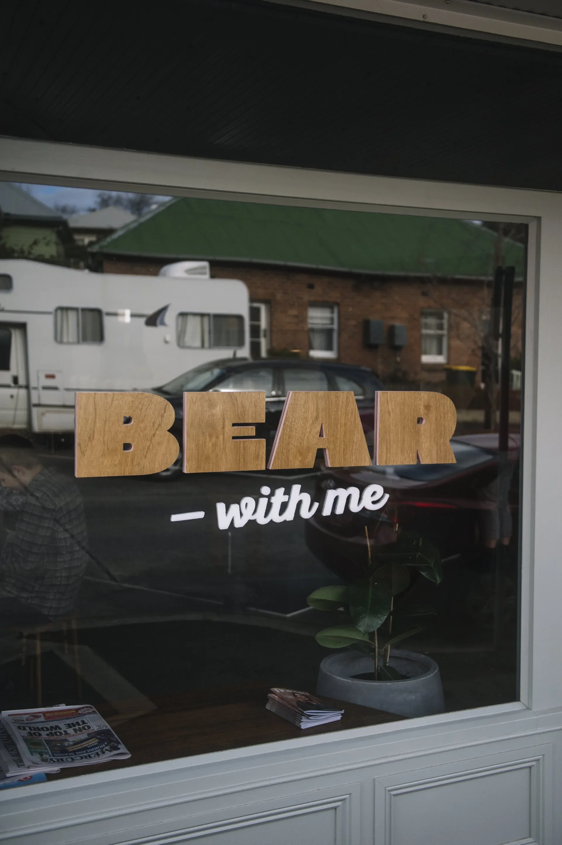 Wooden "BEAR" lettering with white cursive "with me" script is applied to a window, reflecting the streetscape behind, while framing a view into the interior space.