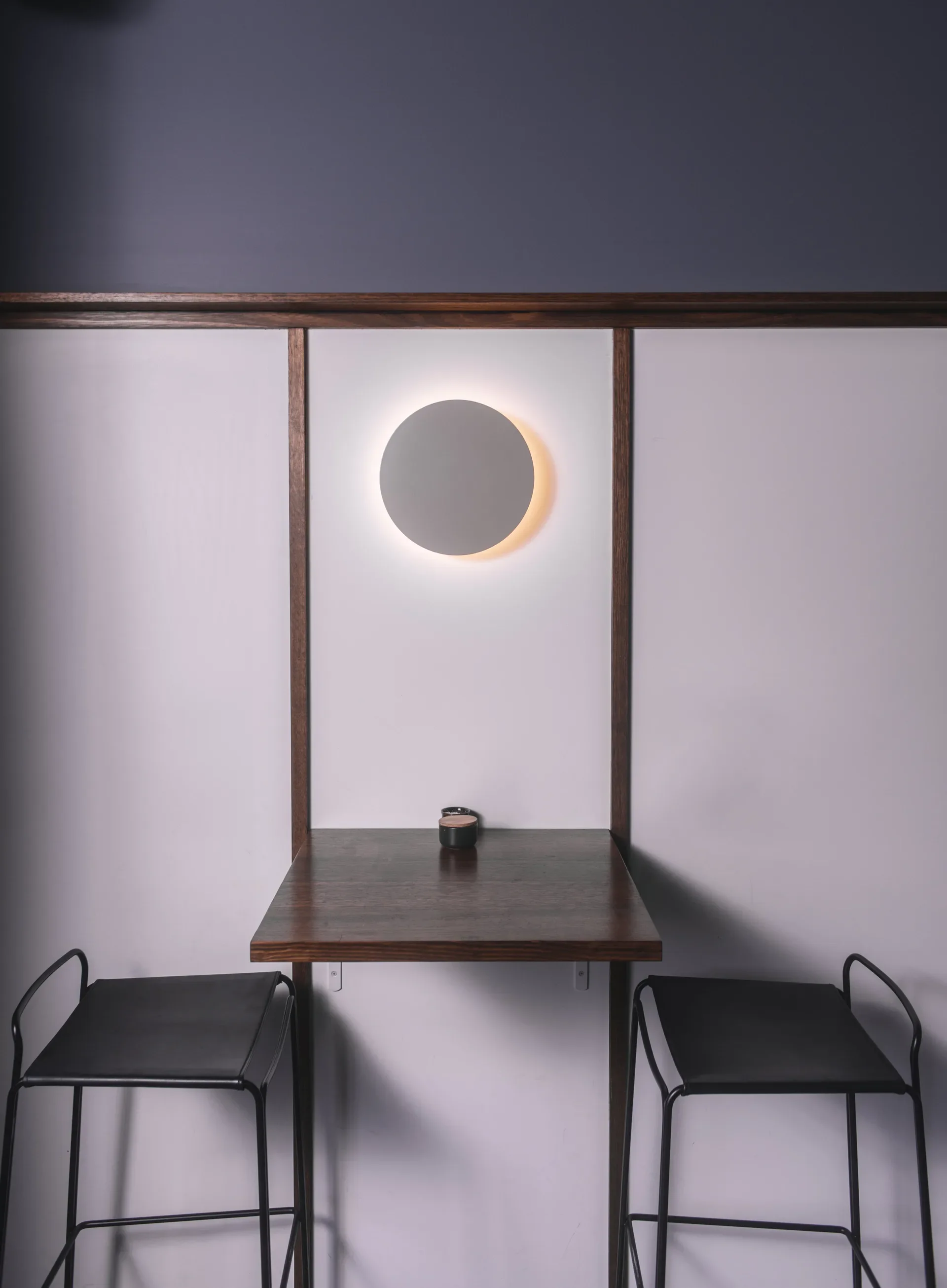 A minimalist seating space features a floating timber table and black wire stools set against a white wall, framed by timber battening and centered under a glowing circular wall light.