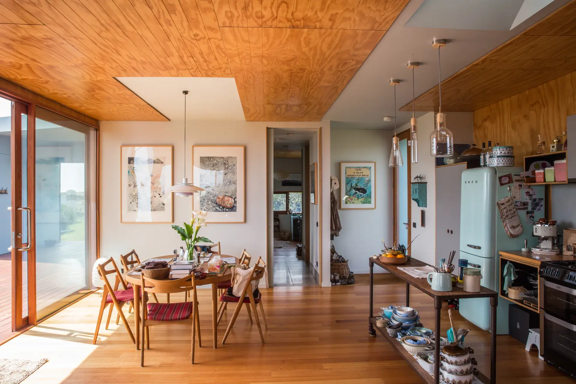 A photograph of the kitchen and dining area, where timber ceilings, warm joinery, and filtered daylight create a relaxed, lived-in interior connected to the outdoors.