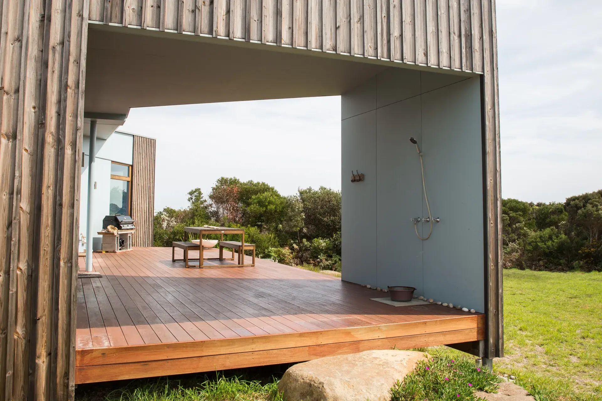A photograph of the covered outdoor deck, complete with outdoor shower and timber decking beneath a framed opening that captures sky and vegetation.