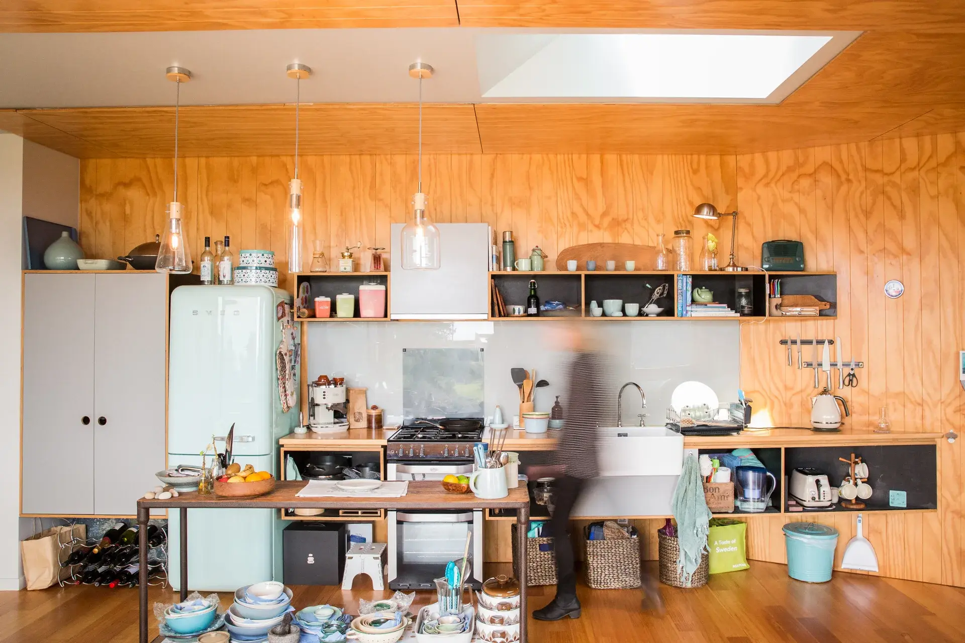 A photograph of the open-plan living area, lined in plywood with integrated shelving, skylights, and large windows that draw in light and frame views of the surrounding bush.