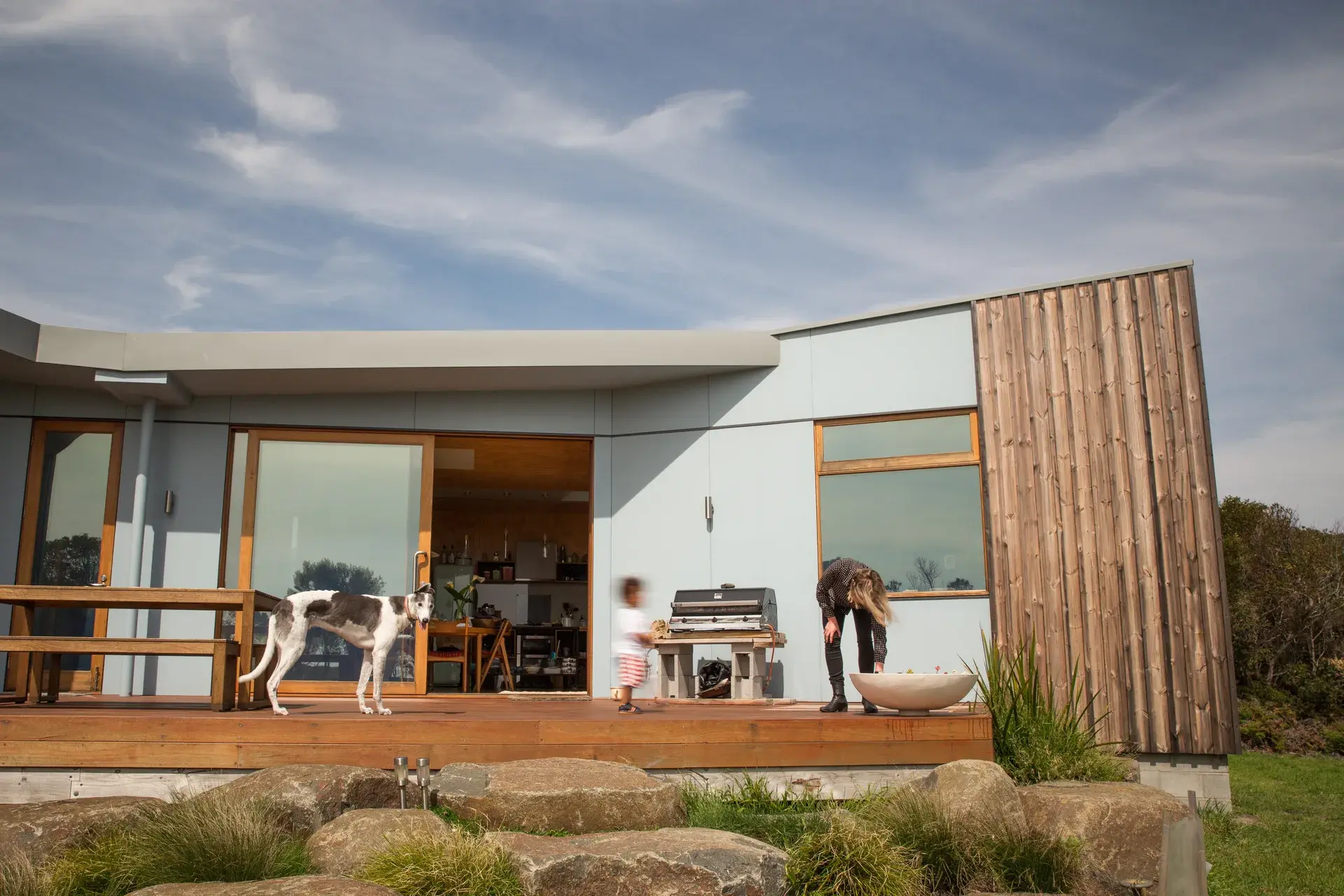 A photograph of the Bell Buoy Beach House house’s low-slung coastal dwelling with timber cladding and pale blue panels, set lightly within a grassy landscape beneath an expansive sky. Large sliding doors open to outdoor living, with a family, dog, and barbecue activating the threshold between interior and landscape.