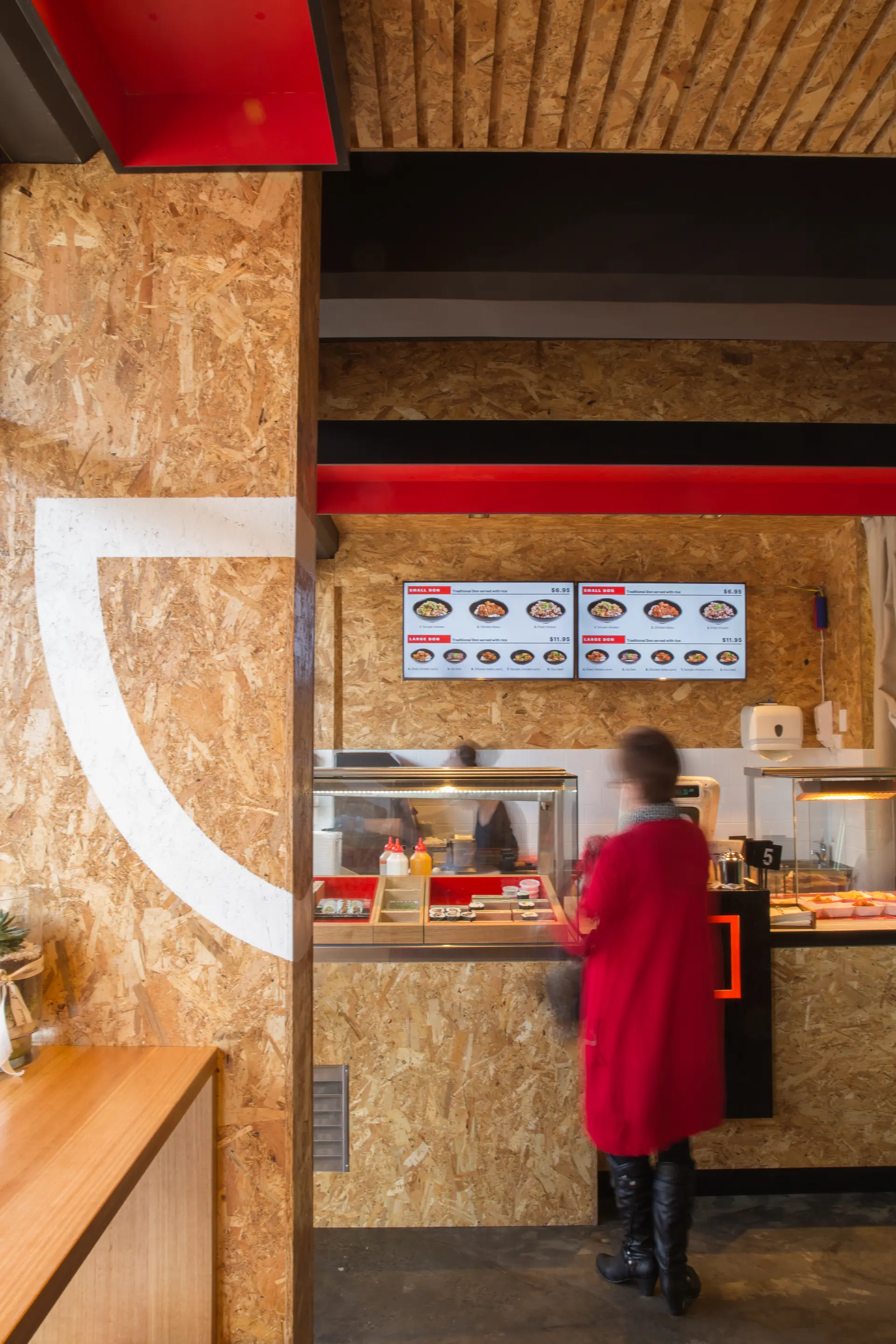A close view of the service counter at Bento Hobart framed by OSB-lined walls and ceiling, with bold red soffit elements above matching the branding palette. A customer stands at the counter, where illuminated menu boards, stainless steel surfaces, and neatly arranged condiments define the compact, efficient ordering space.