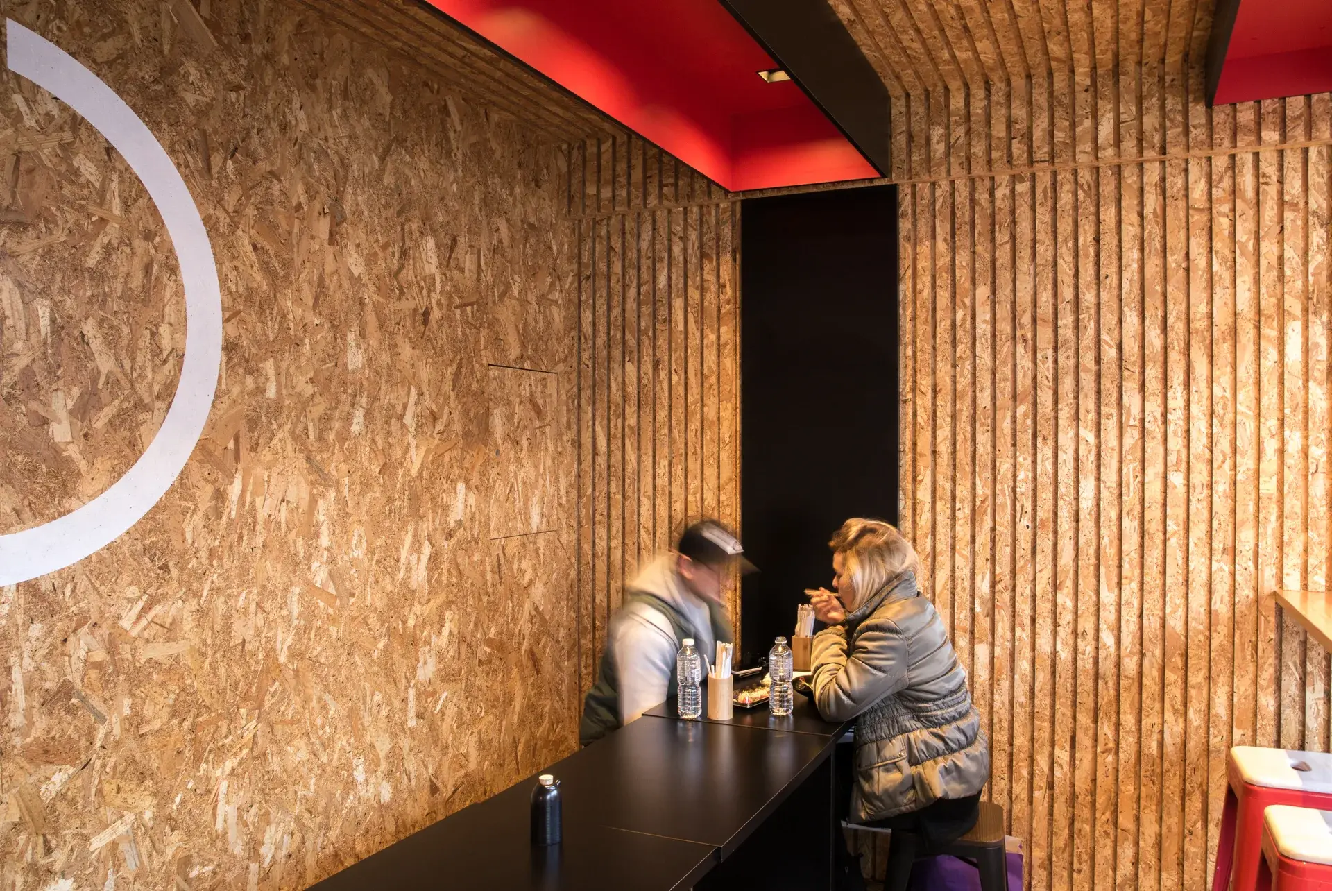 A more intimate seating nook in the corner at Bento Hobart, vertically ribbed OSB panels, where a small but elongated black table with stool seating extends its surface up the adjoining wall and over the ceiling, fitted with an overhead a red-lit ceiling element casts a warm glow in the space during the day.