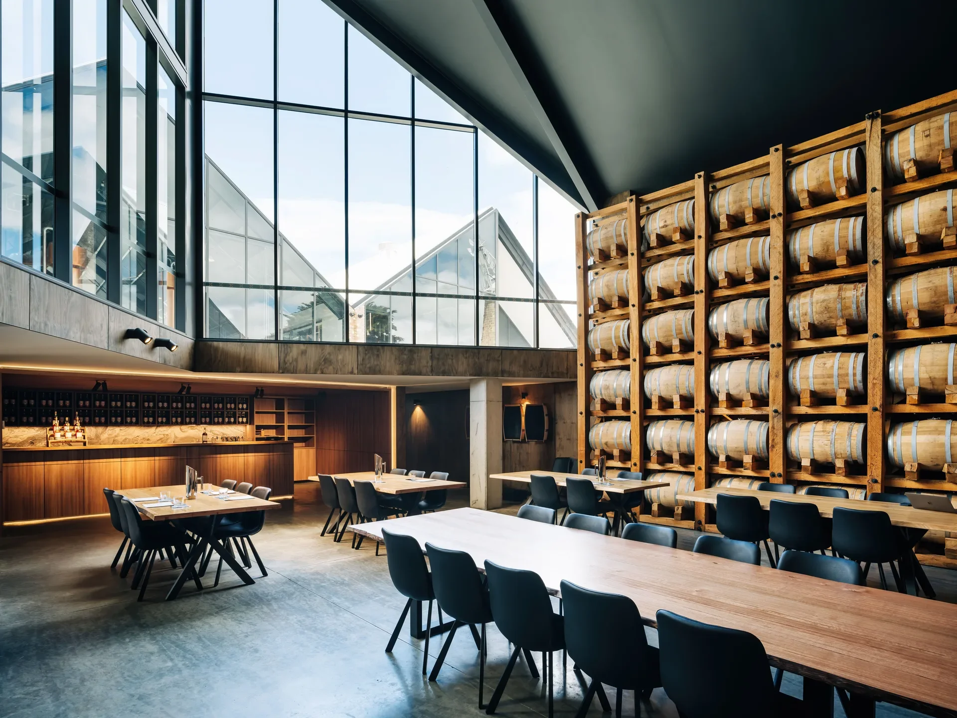 A striking interior featuring a large wall of stacked wooden barrels and clean-lined timber furniture, all housed beneath a high-pitched ceiling and expansive geometric windows.
