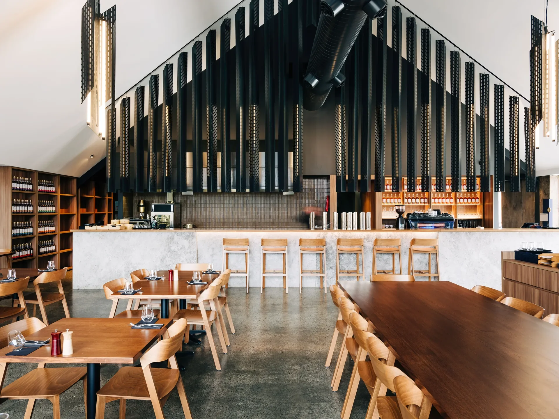 A modern dining hall featuring warm timber tables and chairs, behind a sleek marble-topped bar, all positioned beneath vertical black slats that conform to the shape of the gable roof..