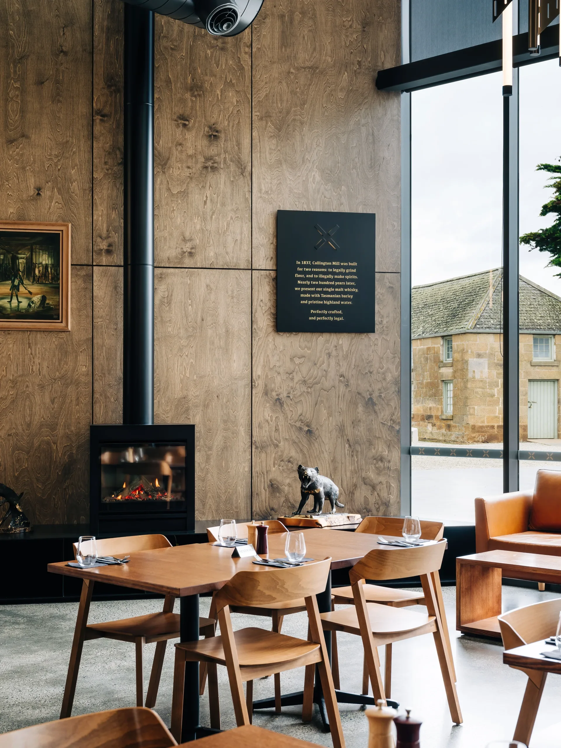 A warm, light-filled dining area features timber tables and chairs set against a wood-paneled wall with a modern fireplace, framed by a large window looking out toward a historic stone building.