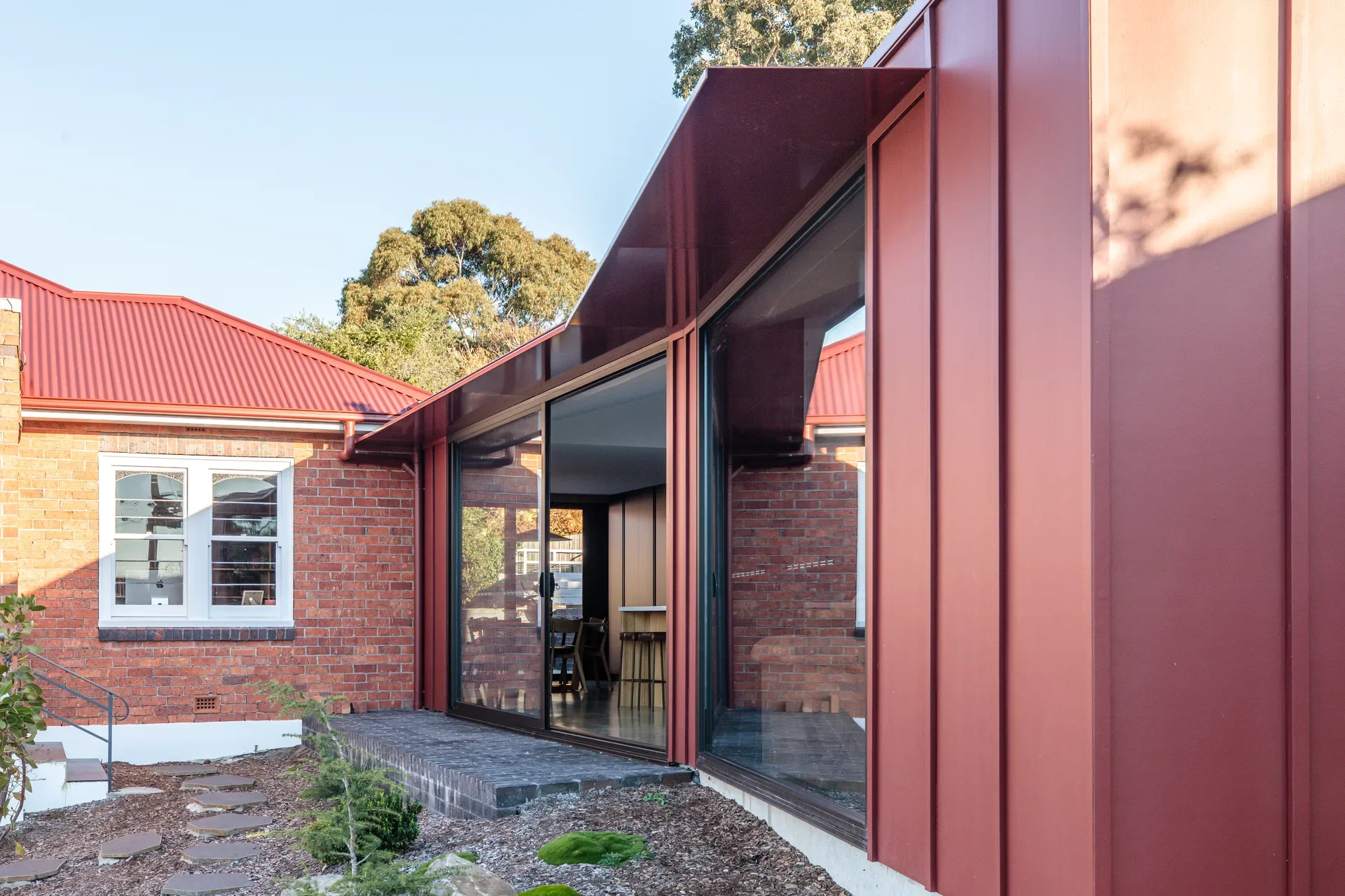 A contemporary red metal-clad extension with a deep overhanging eave and large sliding glass doors creates a bold contrast against the original red brick cottage and traditional gabled roofline.