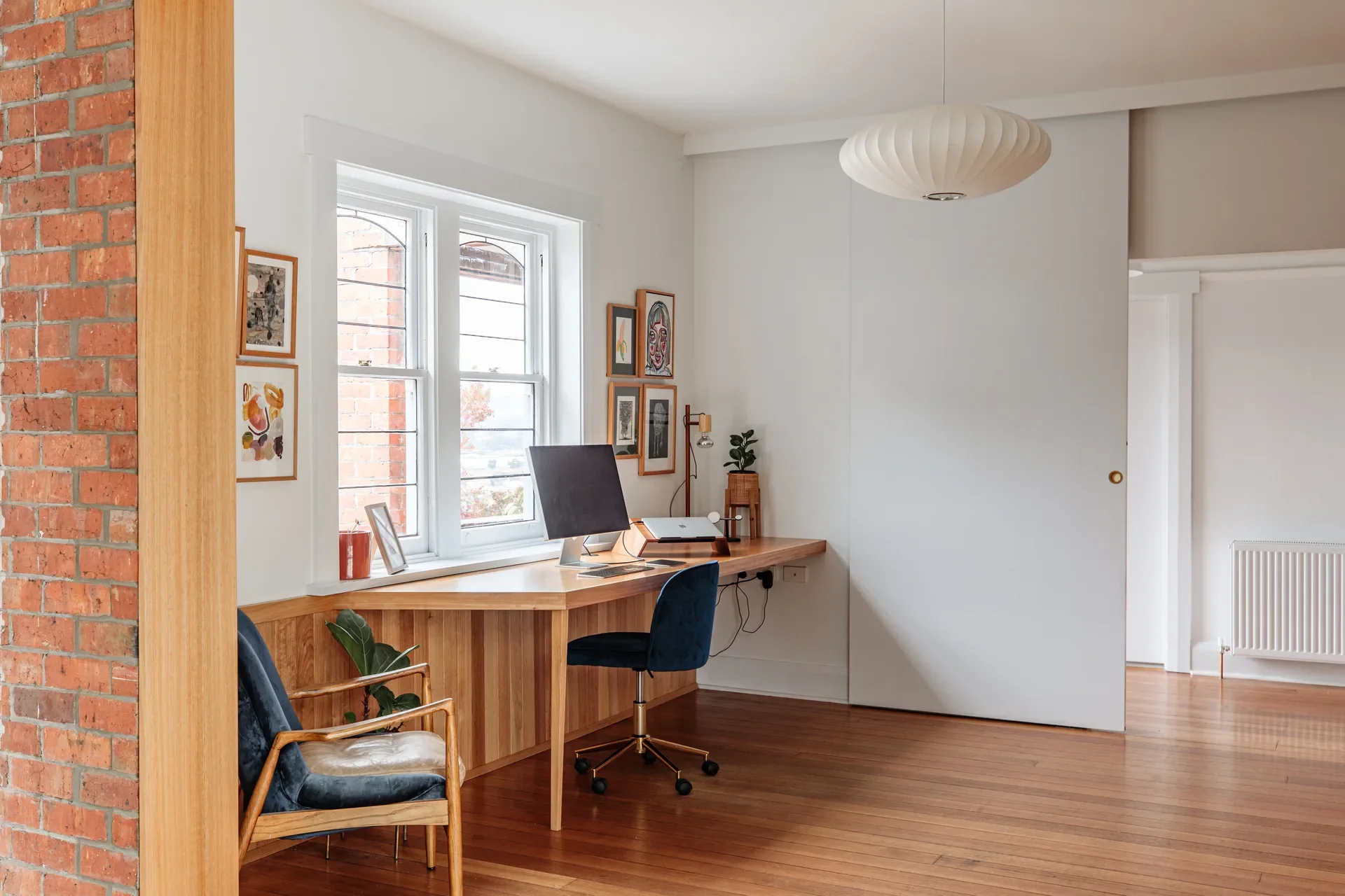 A bright, minimalist study area featuring a long timber desk built under a window, set against white walls, all framed by an exposed brick opening that reveals a polished hardwood flooring.