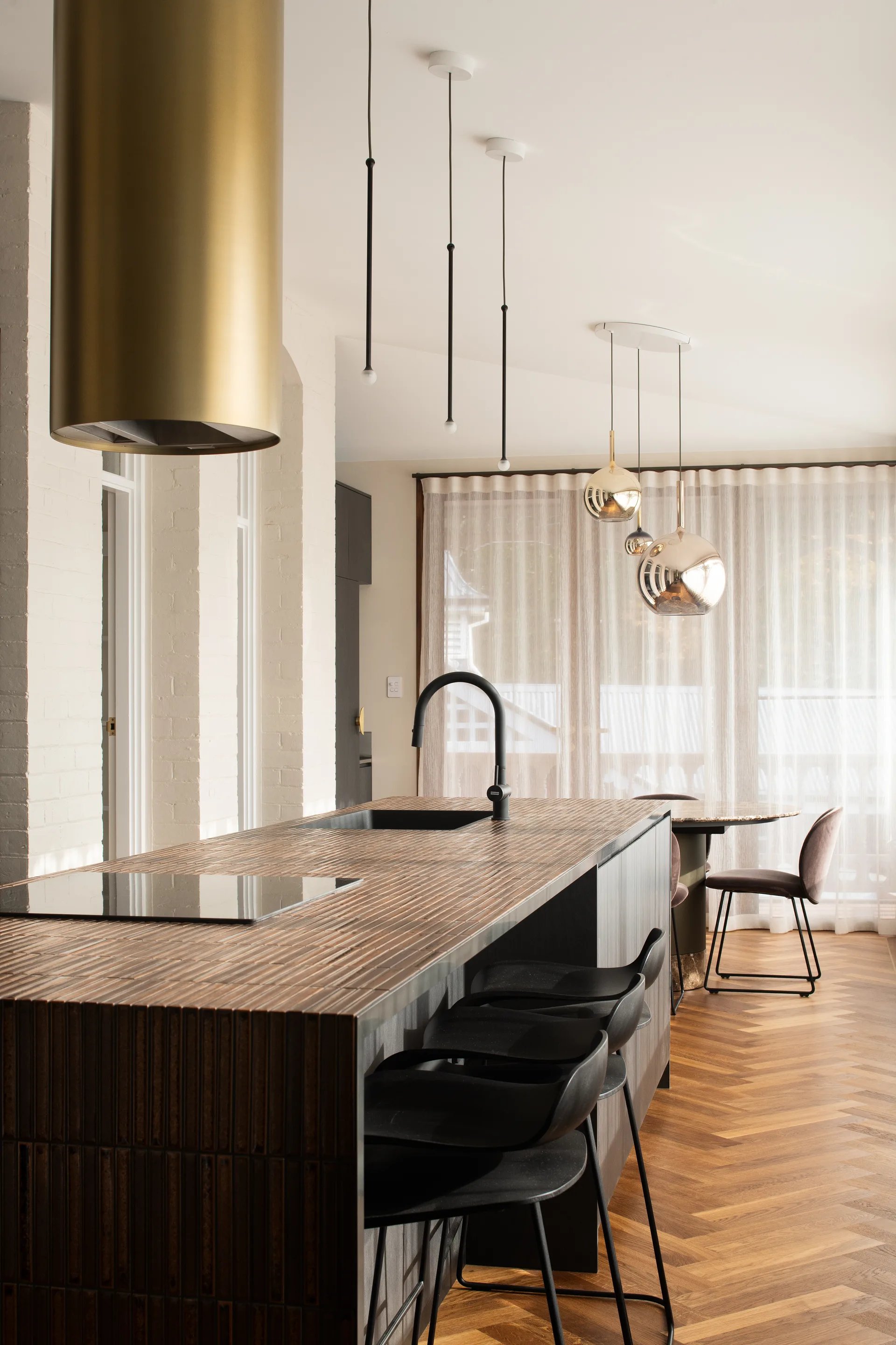 An alternate view of the interior kitchen, looking down the island bench, suspended cylindrical golden flue above the integrated stovetop. A small seating area is at the end, chrome gold pendants against a backdrop of sheer ivory curtains.
