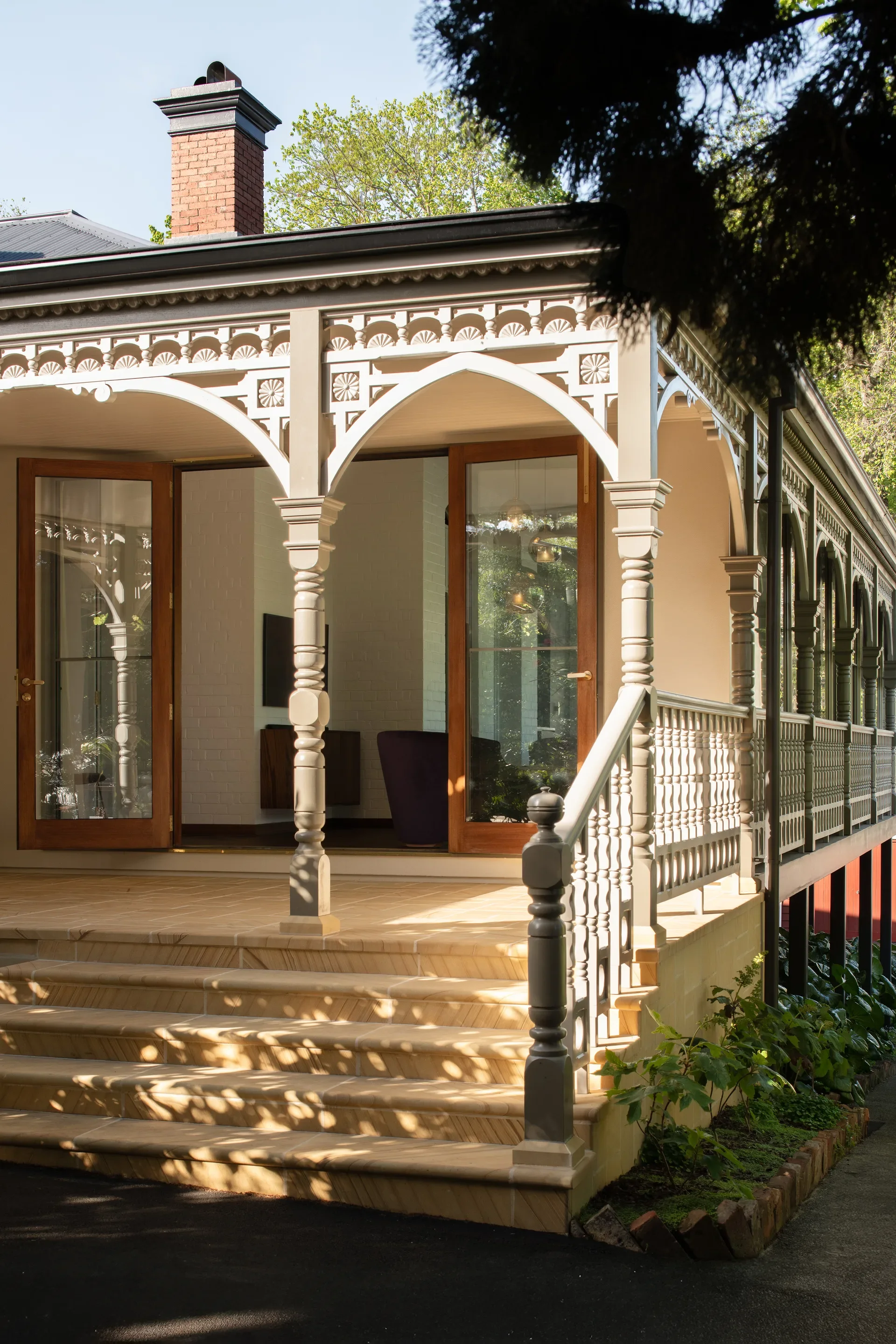 Front entrance to the newly enclosed verandah in dappled light, stone steps lined with the ornate existing timber balustrade and beneath decorative existing arches, leading up to glazed hinge doors with timber framing