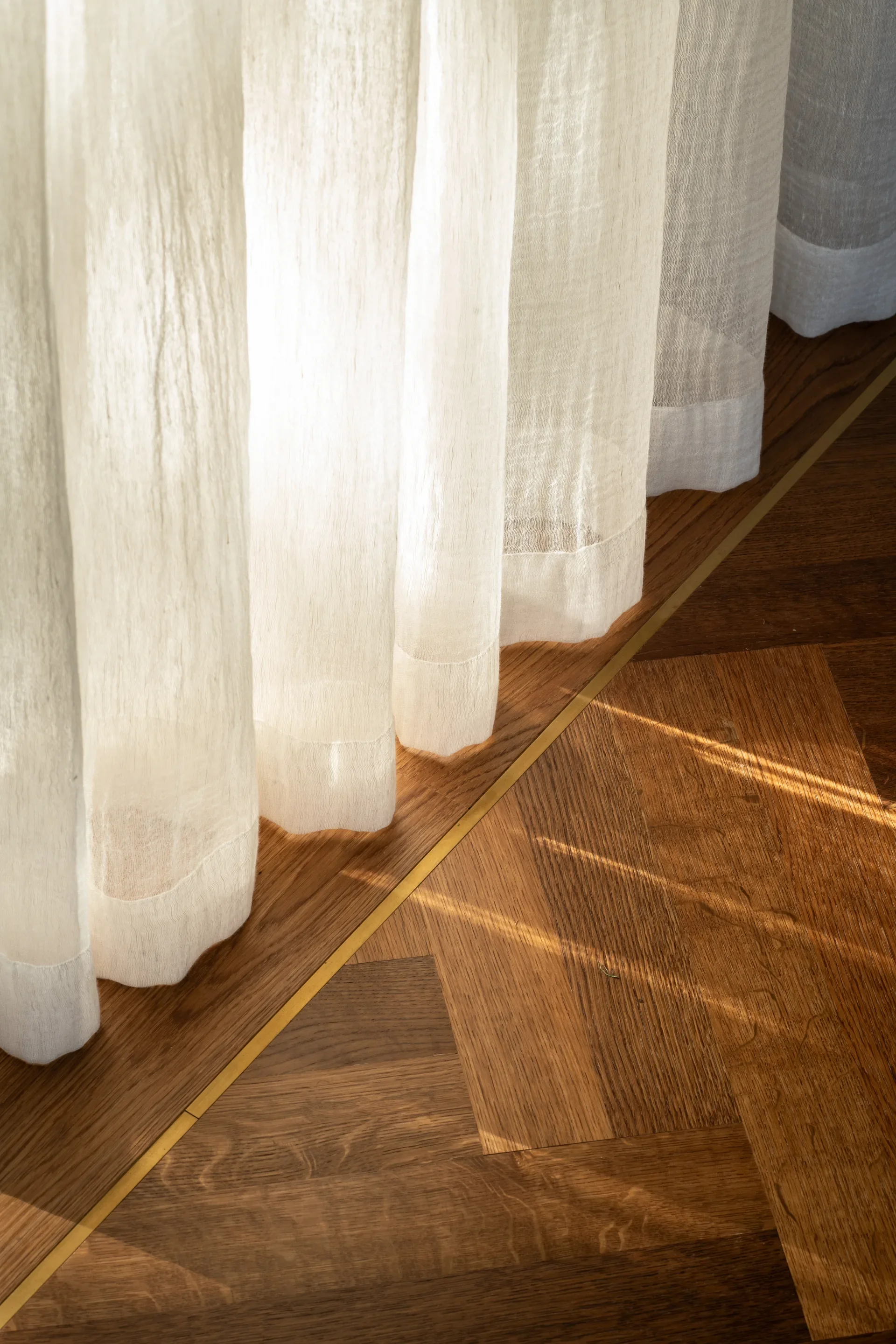 Detail of sheer ivory curtains filtering sunlight over the rich herringbone timber flooring, a brass inlay trim sits at the threshold.