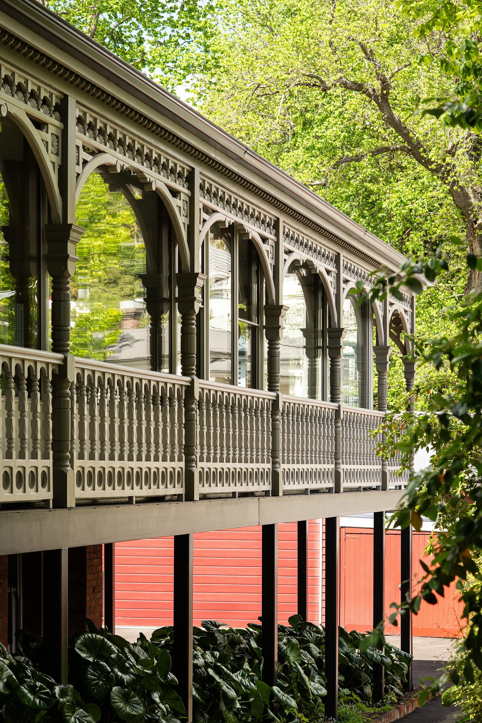 The elevated heritage timber verandah with ornate balustrades, carved posts, and decorative arches retains its original fabric while new full-height glazing is inserted behind the verandah line, creating a layered façade. Framed by mature trees and dense planting, the greenery is reflected across the new glass.