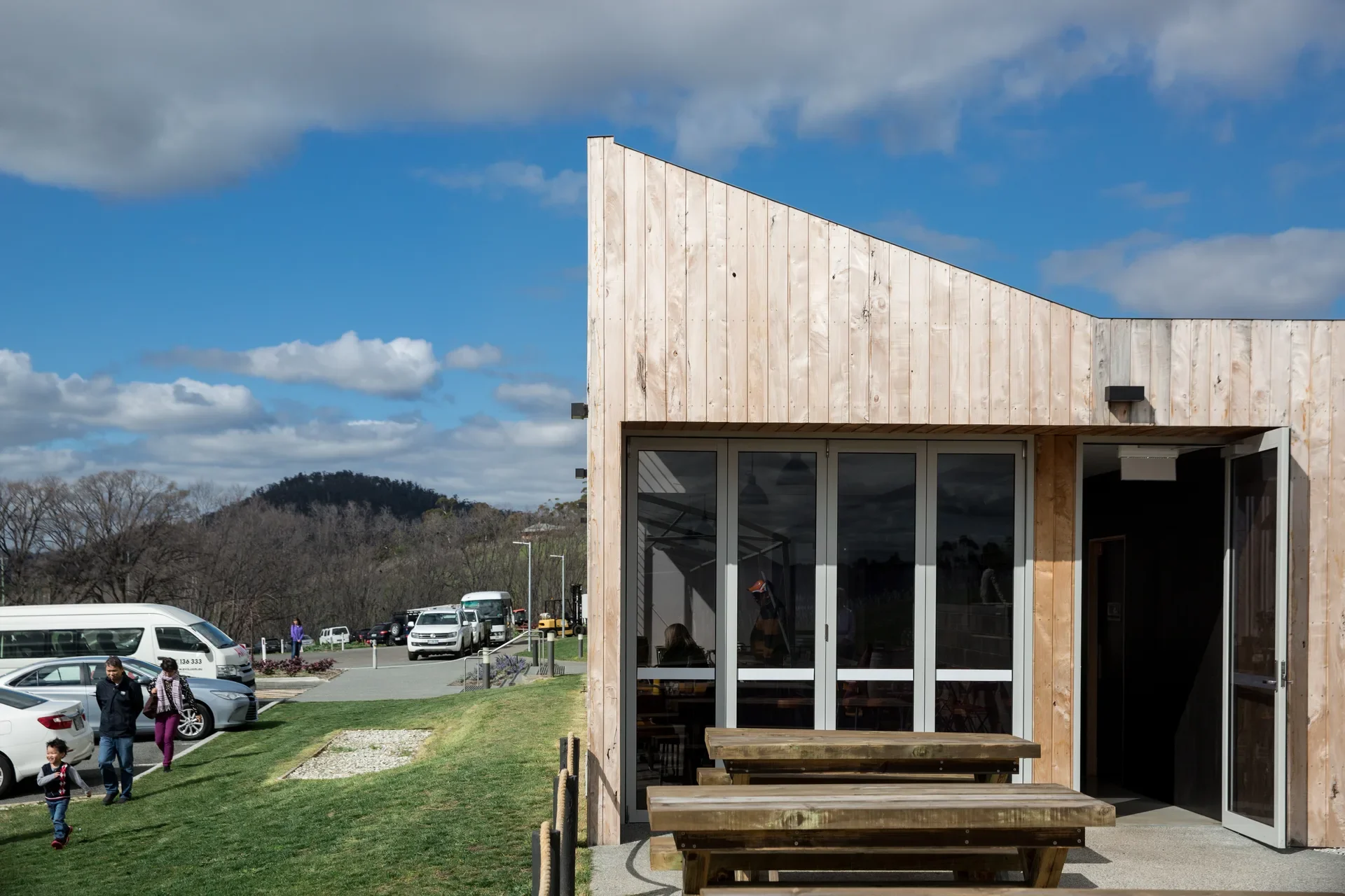The angular side profile of the timber clad building with a bright sky and visitor carpark in the background.