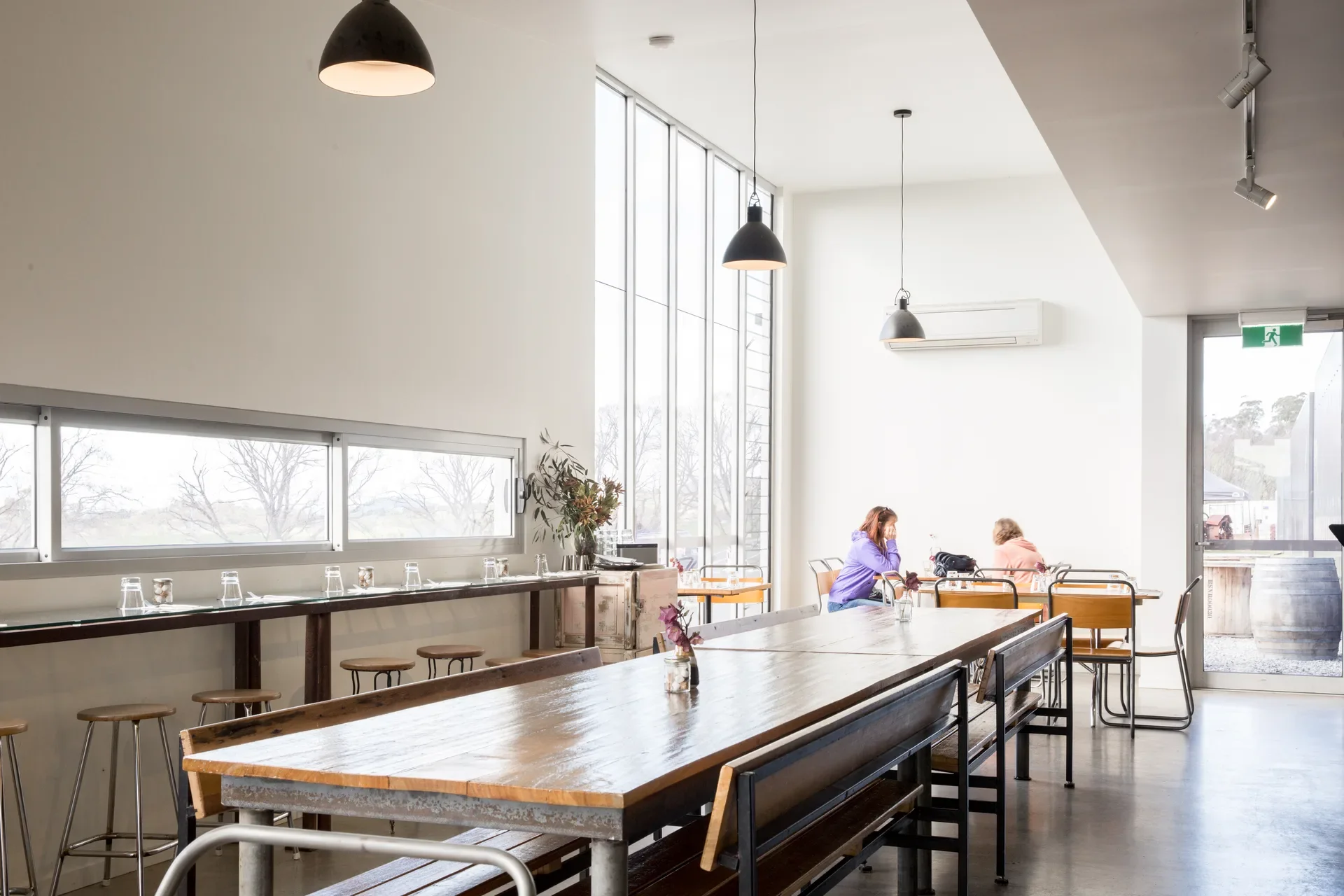 The large commual central table in the light filled cafe space, with smaller tables and architecturally designed windows of varying heights in the background.