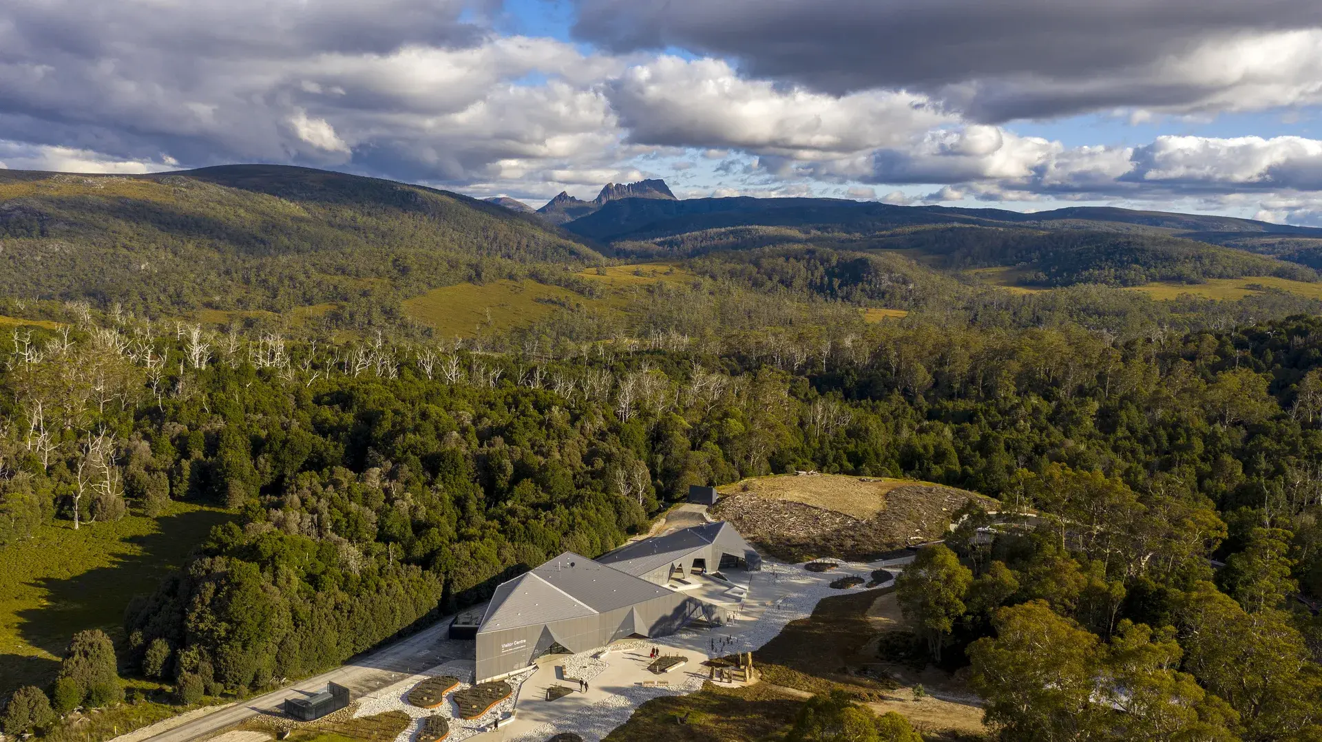 An aerial view reveals the dark, angular visitor centre nestled within a vast expanse of lush forest and rolling hills, set against the backdrop of a distant mountain peak under a dramatic, clouded sky.