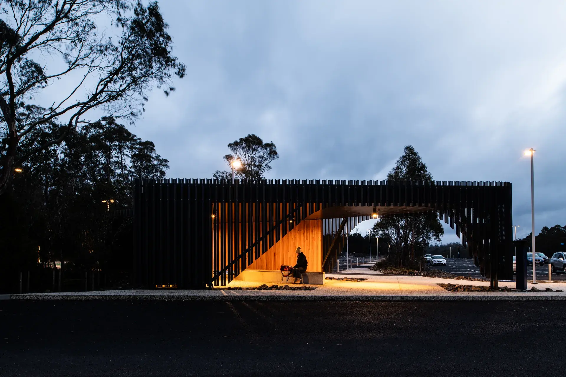 A striking black slatted structure at dusk features a warm, timber-lined alcove with integrated seating, creating a glowing focal point against the darkening trees and overcast sky.