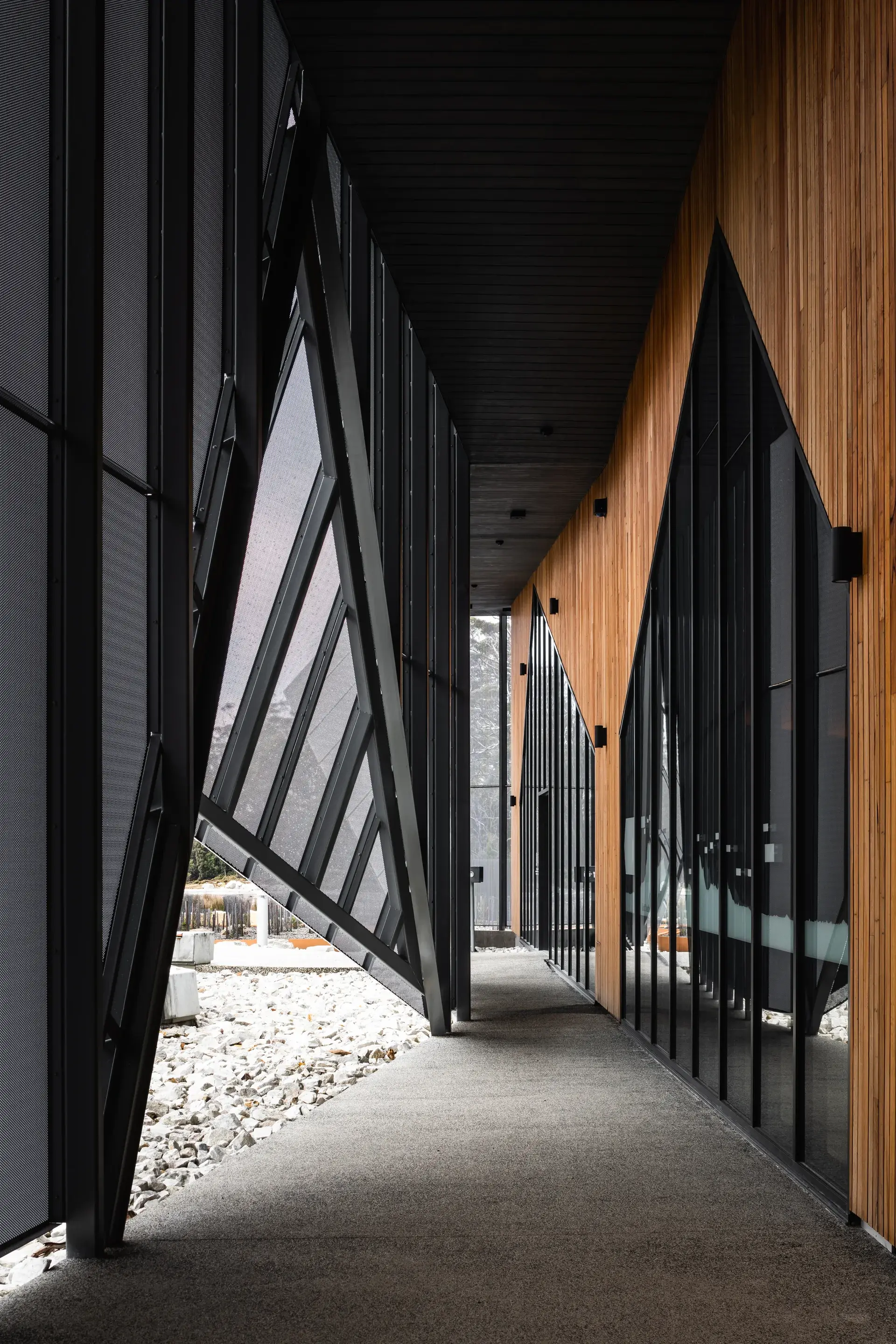 A narrow, covered walkway features a contrast between warm vertical timber paneling and a dark, geometric mesh screen that angles outward toward a rocky landscape.
