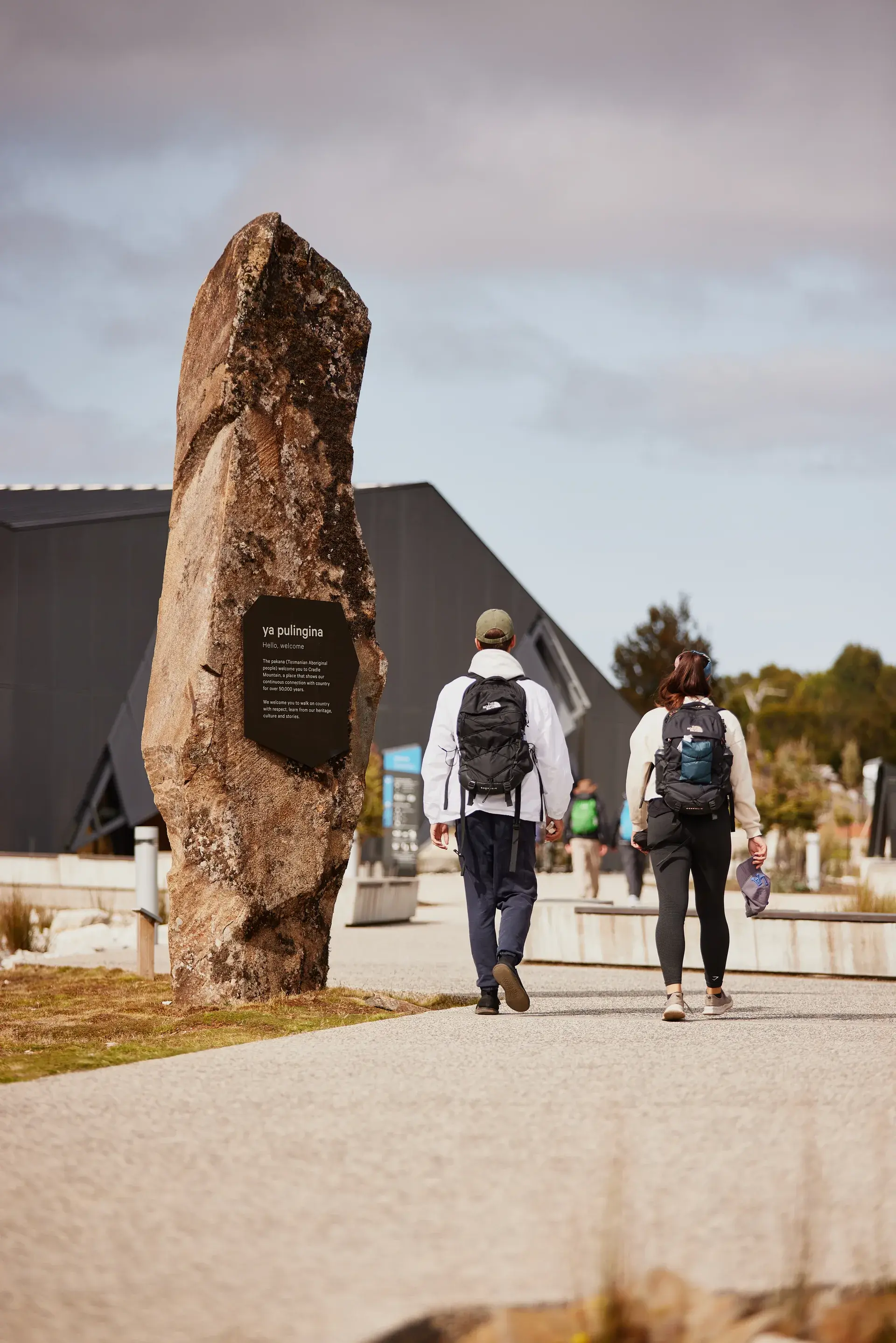 Two hikers with backpacks walk along a gravel path towards the dark geometric building, passing a large, weathered stone monolith inscribed with a "ya pulingina" welcome sign.