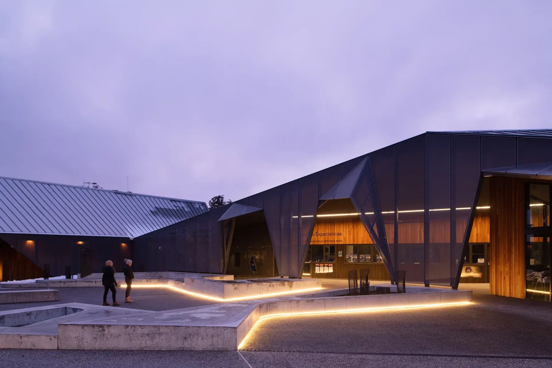 A geometric, dark-panneled building at twilight features sharp, angular entryways and illuminated concrete planters that guide visitors toward the warm, timber-lined interior.